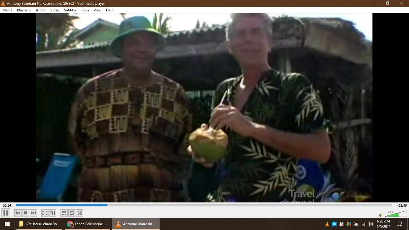 In this frame from the show "Anthony Bourdain: No Reservations," Anthony Bourdain is seen standing outdoors in a casual setting. He is holding a coconut with a straw, suggesting a tropical location. Bourdain is wearing a dark shirt with a green leafy pattern, which complements the laid-back atmosphere. Beside him stands another person, dressed in a patterned shirt and green bucket hat, adding to the cultural and local vibe of the scene. They appear to be engaging in a friendly conversation, possibly discussing local customs or cuisine. The background features a rustic building and some greenery, reinforcing the tropical ambiance. The lighting is bright, indicating daytime, and the overall mood is relaxed and informal, typical of Bourdain's explorative and engaging style. The video is playing on VLC media player, as indicated by the interface visible in the image.