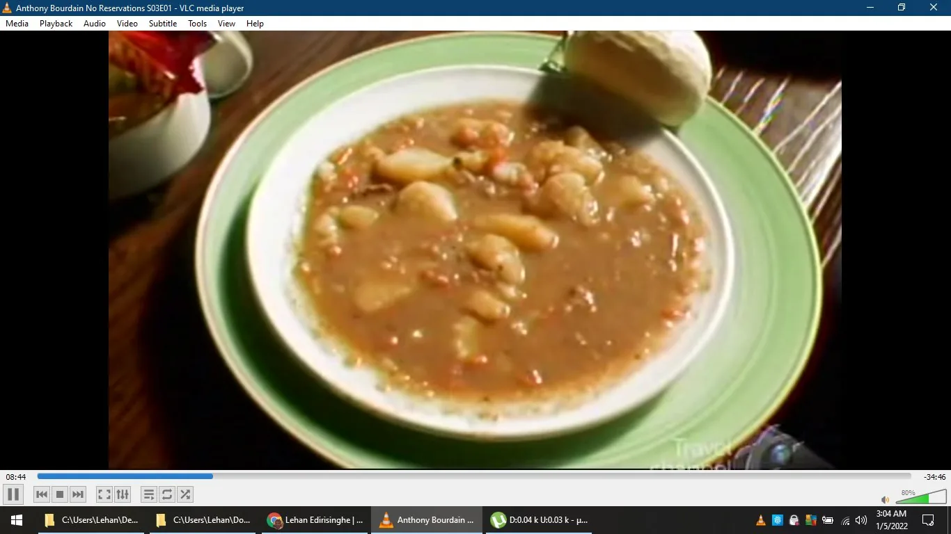 The image is a frame from the show "Anthony Bourdain No Reservations" Season 3, Episode 1, as indicated by the title displayed in the VLC media player window. The scene shows a plate of hearty soup on a table, paired with a piece of bread. The soup appears thick and chunky, featuring pieces of vegetables and possibly beans, suggesting a rustic and comforting dish. The plate is placed on a green-bordered dish, enhancing the warmth of the meal with earthy tones.
The video is playing on VLC media player, with typical interface elements visible, such as the play/pause button, timeline, and volume control. The desktop background reveals open folder paths, suggesting a casual setting or a personal viewing experience. The Travel Channel's logo is faintly visible in the bottom right corner, confirming the network associated with the show. The overall lighting is warm, emphasizing the inviting nature of the meal, reflecting the travel and culinary theme of the series hosted by Anthony Bourdain.
- Title: "Anthony Bourdain No Reservations S03E01"
- Site / app: "VLC media player"