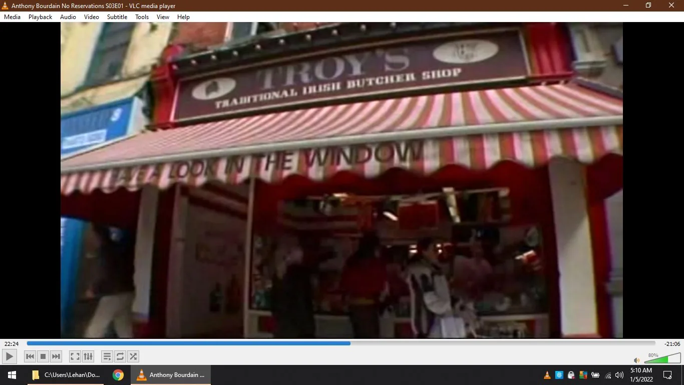 ```
**SECTION 2: Rich prose with relevant info**
This image is a frame from the video "Anthony Bourdain No Reservations S03E01," viewed using VLC media player. The scene captures the exterior of Troy's Traditional Irish Butcher Shop, recognizable by its striped red and white awning and a sign that reads "LOOK IN THE WINDOW." The setting appears to be outdoors on a bustling street, likely in an Irish town given the theme of the shop. The awning provides a quaint, traditional aesthetic to the front of the shop.
In the foreground, a few people are visible, standing or walking near the shop entrance, dressed casually, indicating a lively and approachable atmosphere typical of a busy market area. The shop seems to be well-visited, with an inviting entrance that encourages passersby to explore inside. The lighting suggests it is daytime, casting bright, natural light over the scene. This frame highlights the cultural and culinary exploration theme central to Anthony Bourdain's show, where he often delves into local traditions and food cultures.