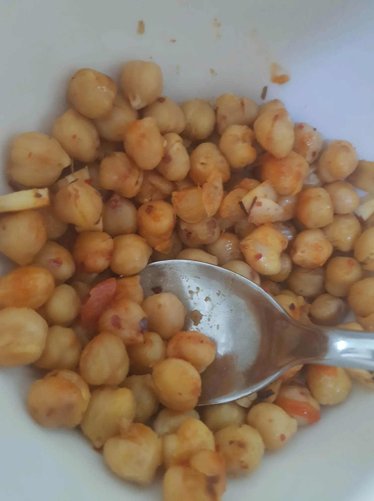 This image shows a close-up of a bowl filled with seasoned chickpeas. The chickpeas appear to be cooked and are seasoned with what looks like red chili flakes or similar spices, giving them a slightly reddish hue in some areas. There is a metal spoon partially visible, suggesting the dish is ready to be served or mixed. The lighting is bright, likely natural or from an indoor source, highlighting the texture and color of the chickpeas. The bowl itself is white, providing a neutral background that contrasts with the colorful dish. The overall setting suggests a casual, home-cooked meal preparation.