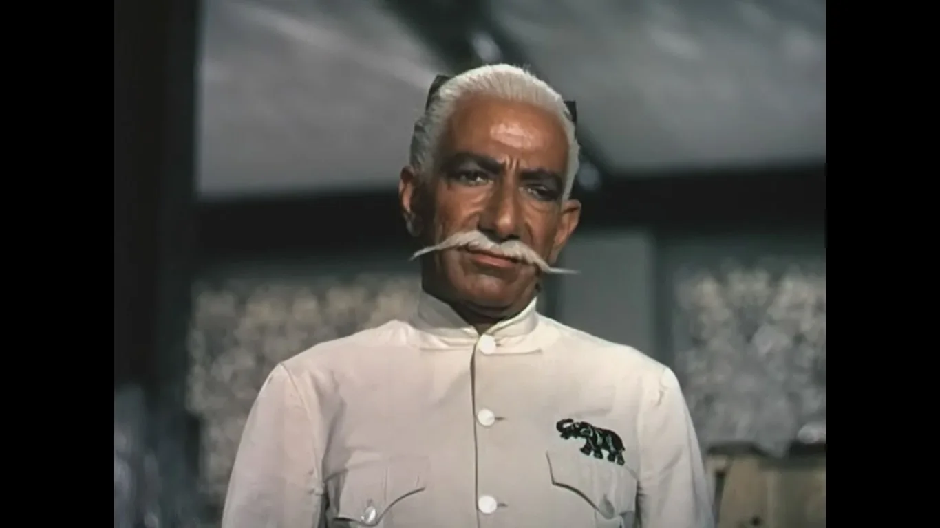 The image depicts a serious character from the 1954 film "Elephant Walk." The setting appears to be indoor, with a subdued atmosphere. The character is an older man with a distinctive white mustache and hair, conveying a sense of authority and dignity. He is wearing a traditional outfit, which is white with a high collar and features a black embroidered design of an elephant on the chest. The expression on his face is stern, suggesting a moment of contemplation or decision. The lighting is soft and focused, accentuating his facial features and the detailed embroidery on his attire. The background is slightly blurred, putting the focus on the character, and it hints at a setting rich in cultural or historical significance.