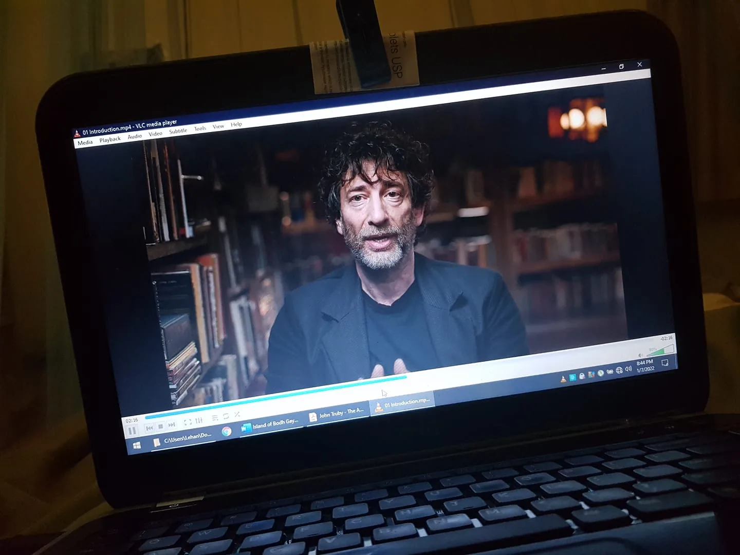 The image displays a laptop screen playing a video in VLC media player. The file name "01 Introduction.mp4" is visible in the title bar. The scene features a middle-aged man with curly hair and a beard, dressed in a dark jacket, sitting in a room that resembles a library or study. Books are visible on shelves in the background, suggesting an intellectual or literary setting. The lighting is warm and subdued, focusing on the man's face and upper body. The video playback bar shows the video is approximately two minutes and sixteen seconds into the introduction, and the current time on the device reads 8:41 PM. The expression of the man appears thoughtful, likely indicating he is explaining or discussing a topic. The laptop screen setup suggests a casual viewing environment, possibly for educational or personal enrichment purposes.