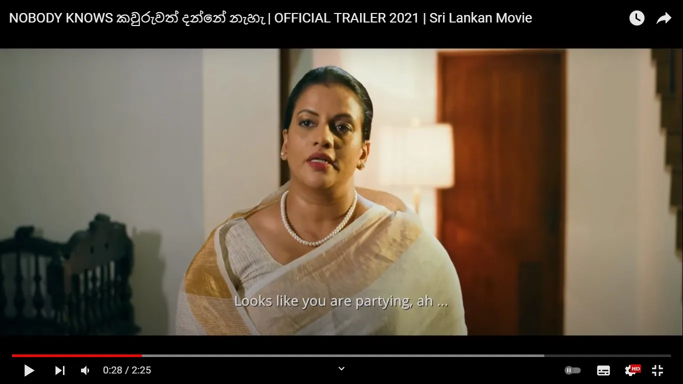 The image is a video frame from the official trailer of the 2021 Sri Lankan movie titled "NOBODY KNOWS කවුරුවත් දන්නේ නැහැ." The scene features Sangeetha Weeraratne, as identified by the user hint. She is indoors, possibly in a domestic setting, as suggested by the wooden chair and lamp in the background. Sangeetha is dressed in a traditional saree with a pearl necklace, indicating a formal or cultural context. Her expression appears serious or inquisitive, fitting for a conversation or confrontation. The lighting is warm and soft, creating an intimate atmosphere. The dialogue subtitle reads, "Looks like you are partying, ah ...," suggesting a casual or slightly confrontational remark. The video is hosted on YouTube, and the frame is timestamped at 0:28 of a 2:25-minute trailer.