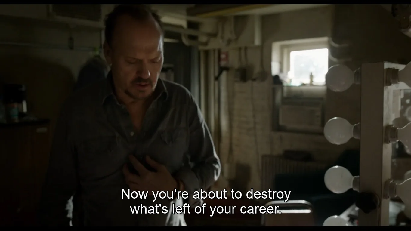 In this scene from the film "Birdman," Michael Keaton's character is captured in a tense moment inside what appears to be a dimly lit dressing room. The lighting casts shadows, creating a somber atmosphere that matches the serious tone of the situation. Keaton, wearing a dark gray button-up shirt, appears deep in thought, his hand placed on his chest as if contemplating or feeling stressed. The room is cluttered, with a makeup mirror lined with light bulbs visible on the right side of the frame, suggesting a theater setting. In the background, there is an air conditioning unit under a small window, adding to the room's utilitarian feel. The subtitle reads, "Now you're about to destroy what's left of your career," indicating a critical and introspective moment for the character. The entire composition and mood reflect themes of self-reflection and the pressures of maintaining a career in the arts.