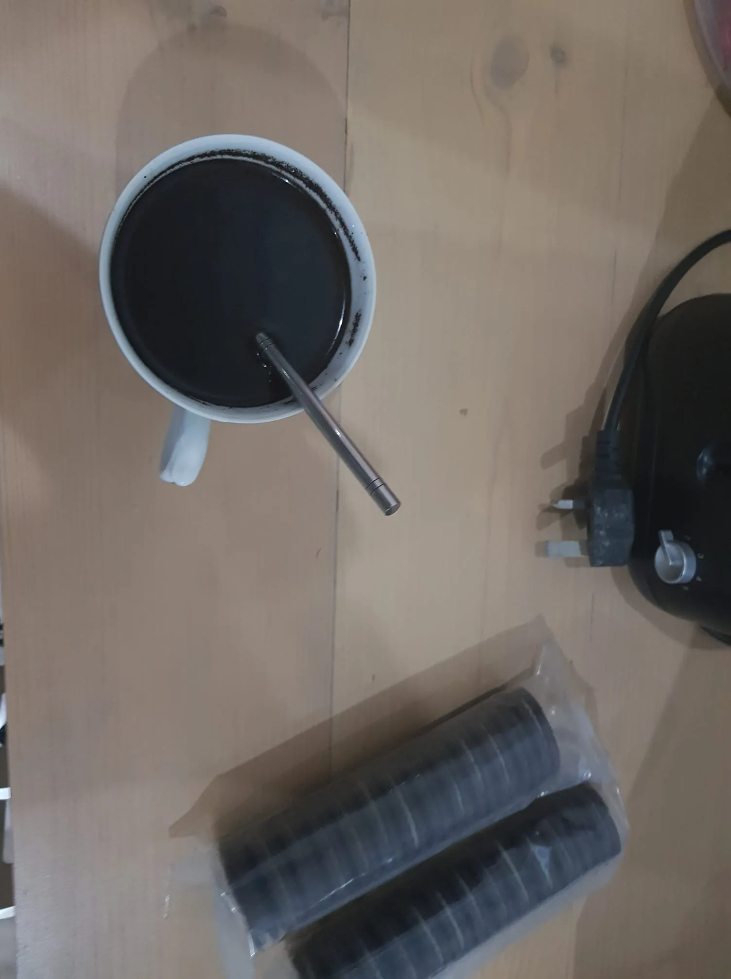 The image shows a wooden table with several items on its surface. In the center, there is a white cup filled with a dark liquid, likely coffee, with a silver spoon resting inside. The cup's handle is visible on the left side. Adjacent to the cup, two plastic-wrapped stacks of chocolate sandwich cookies are placed, suggesting a pairing with the coffee for a snack. To the right, there is an electronic device with a visible three-pronged plug and a dial, possibly a kitchen appliance or a computer accessory. The lighting is natural and soft, creating a cozy atmosphere. The setting appears to be indoors, possibly a kitchen or dining area. The arrangement hints at a casual moment of relaxation or a quick break.