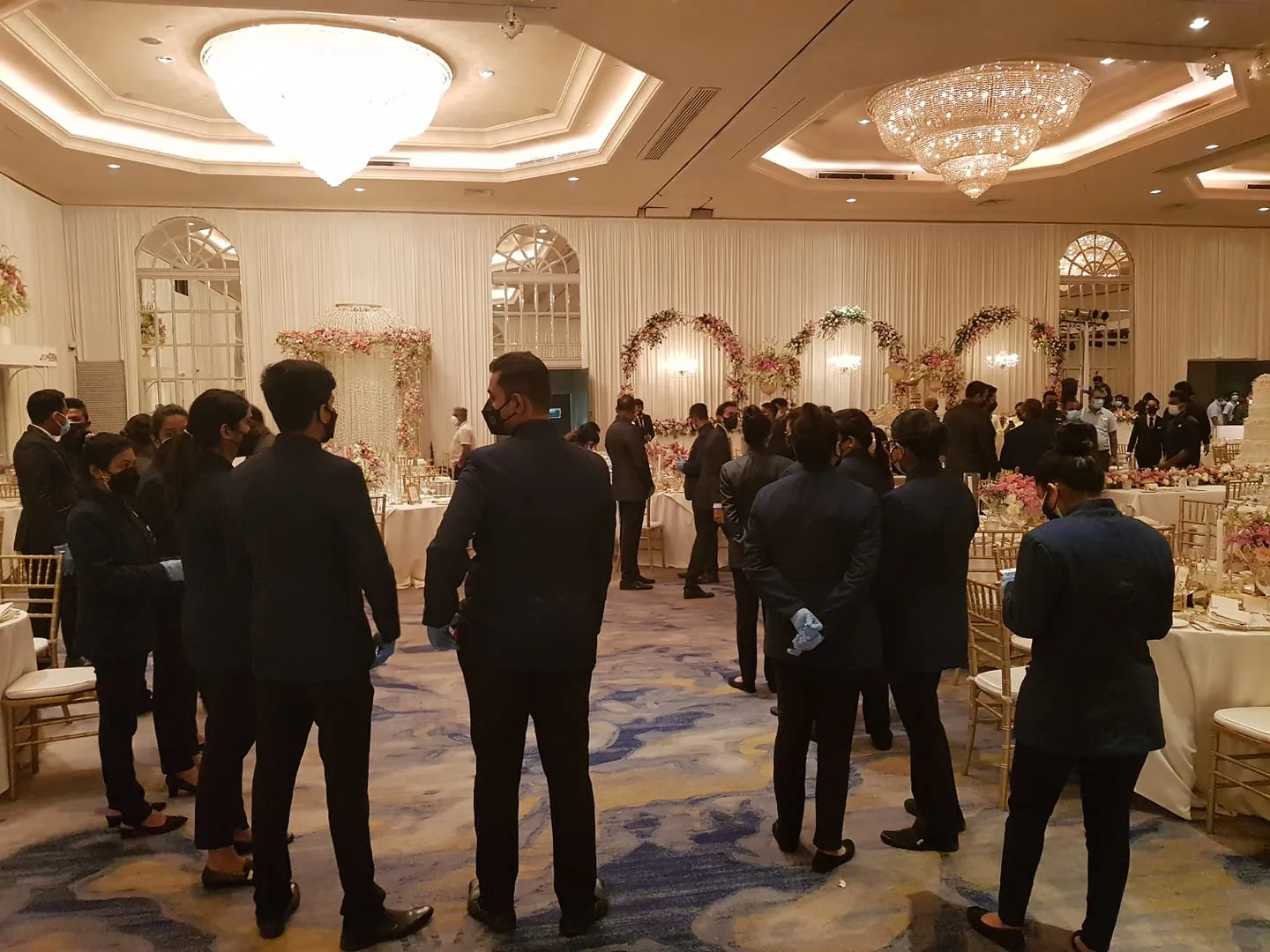 The image depicts an indoor event taking place in an elegant, well-lit banquet hall. The room features large, ornate chandeliers hanging from the ceiling, casting warm light over the space. A group of individuals, likely event staff, are gathered in the foreground, wearing formal black attire and black face masks, indicating a focus on safety protocols. Some staff members also wear gloves, suggesting they are preparing to serve or assist guests.
In the background, tables are set with fine dining arrangements, including floral centerpieces with pink and white flowers, adding a touch of sophistication. Arched mirrors and draped curtains enhance the room's elegance. The overall atmosphere is formal and organized, with the staff appearing attentive and professional, likely preparing for or overseeing a significant event such as a wedding or corporate gathering. The room's design and decor reflect a classic and luxurious style.