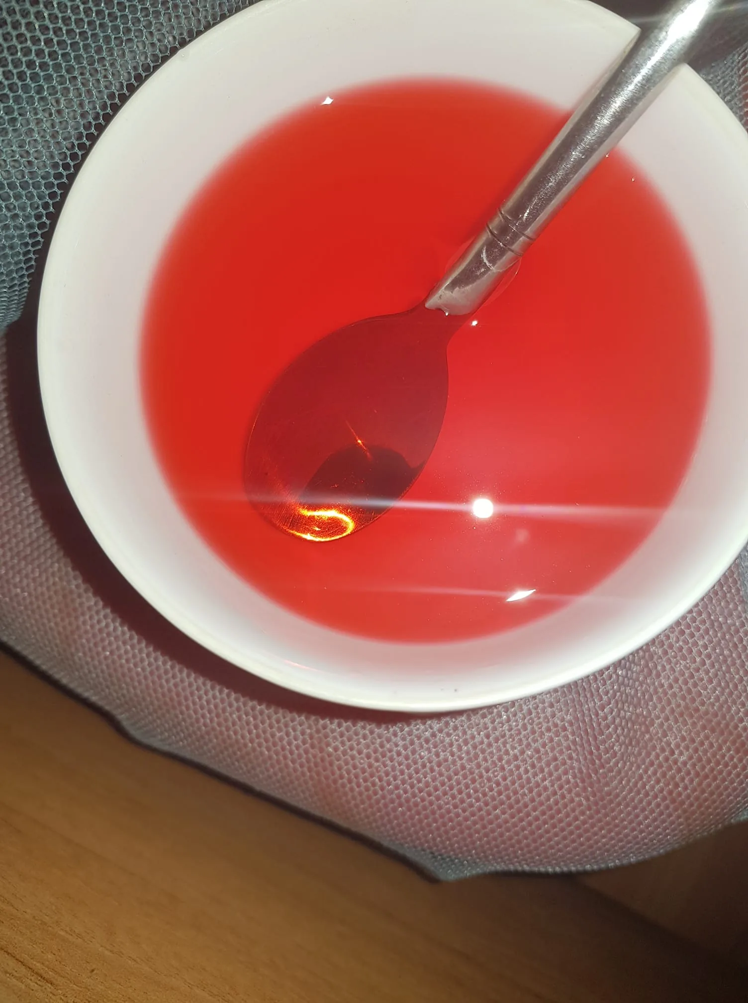The image shows a close-up view of a white bowl containing a vibrant red liquid, possibly a type of drink or soup. Inside the bowl, a silver spoon is partially submerged, reflecting the light above and creating a bright spot on its surface. The liquid appears smooth, with no visible particulates or bubbles, suggesting it is clear. The bowl is resting on a textured, possibly fabric surface, which might be a piece of clothing or a tablecloth. The lighting is bright, casting distinct reflections on the liquid and highlighting its glossy texture. The overall mood is simple and focused, with the red liquid providing a striking contrast against the white bowl and silver spoon. The setting appears to be indoors, with the use of flash or bright artificial lighting evident in the reflections.