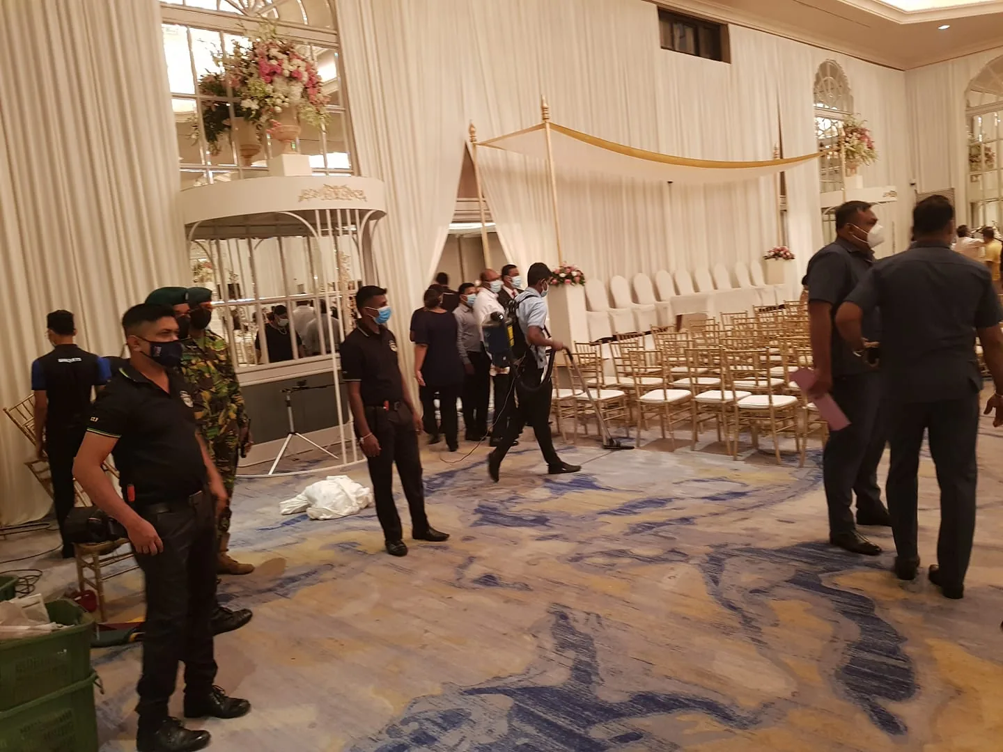 The image depicts an indoor event setup, possibly for a formal occasion. The setting features a large, ornate room with high ceilings and large windows draped with white curtains. In the foreground, several security personnel dressed in uniforms and face masks stand alert, suggesting that preparations are underway or that the event is being secured. Some of them are in black uniforms while one wears camouflage, indicating a mix of security roles.
In the background, rows of gold-colored chairs are neatly arranged facing a raised platform with light-colored upholstery, perhaps for a ceremony or presentation. Floral arrangements accentuate the elegant decor, placed on stands and the platform. The room's lighting is warm, contributing to a formal yet inviting atmosphere. Despite the presence of security, the mood seems calm and organized, indicating preparation or a transition phase in the event timeline. The image captures a blend of elegance and security diligence, hinting at a high-profile or significant gathering.