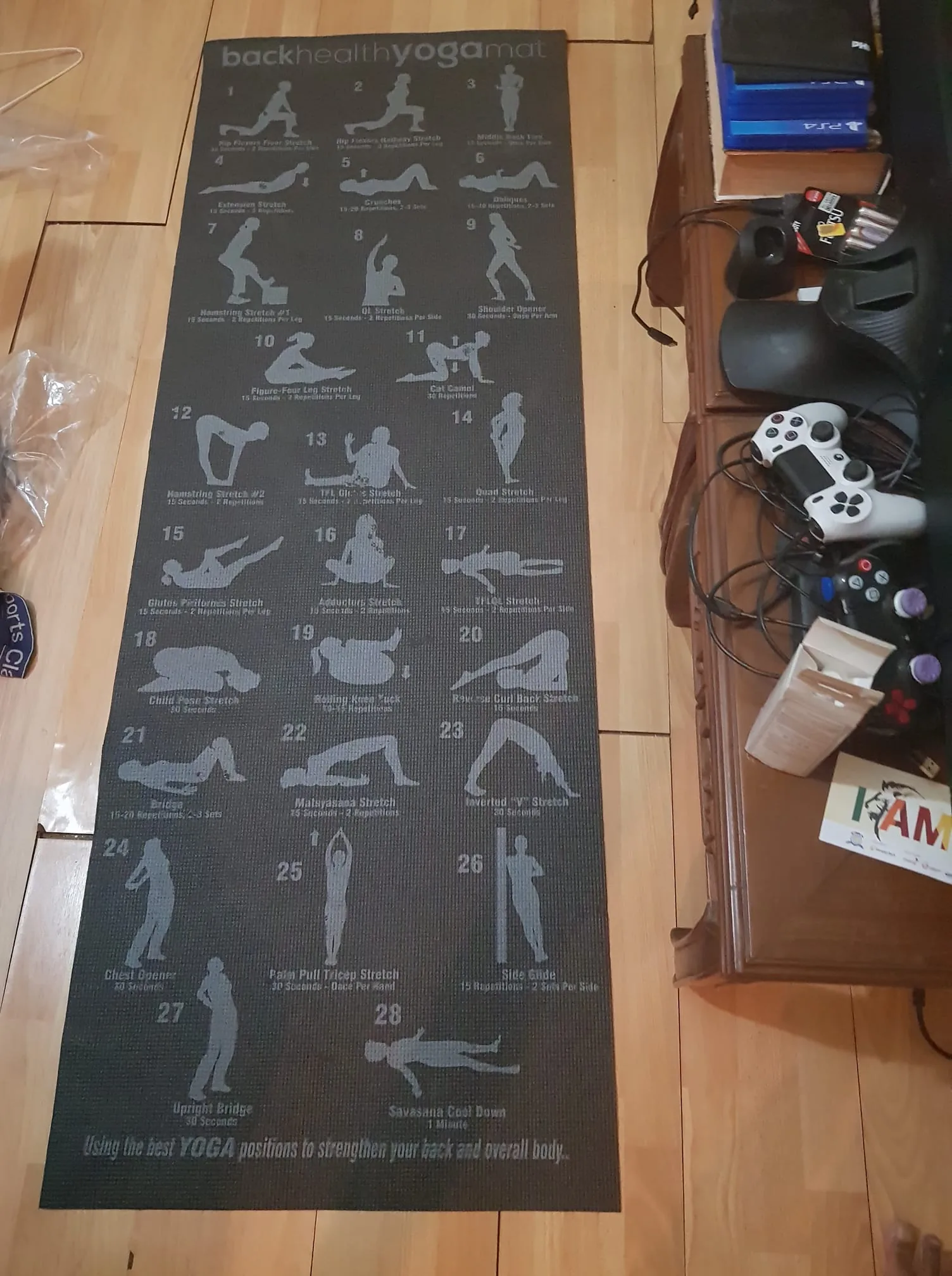 The image shows a yoga mat laid out on a wooden floor, designed with illustrations of various yoga positions. Each position is numbered and labeled with its name and recommended duration, illustrating exercises for flexibility and strength. The mat is titled "back healthy yoga mat," suggesting a focus on exercises to improve back health. The mat's surface is dark with light-colored illustrations and text for contrast, making the instructions easy to read.
On the left side of the image, there is a transparent plastic bag, possibly the packaging for the mat. To the right, the edge of a wooden table is visible, cluttered with video game controllers, a headset, and stacked boxes. This setting suggests a casual living space, perhaps a living room. The lighting is warm, indicating a cozy environment conducive to home workouts.
- Title: "back healthy yoga mat"
- Key Text: "Using the best YOGA positions to strengthen your back and overall body."