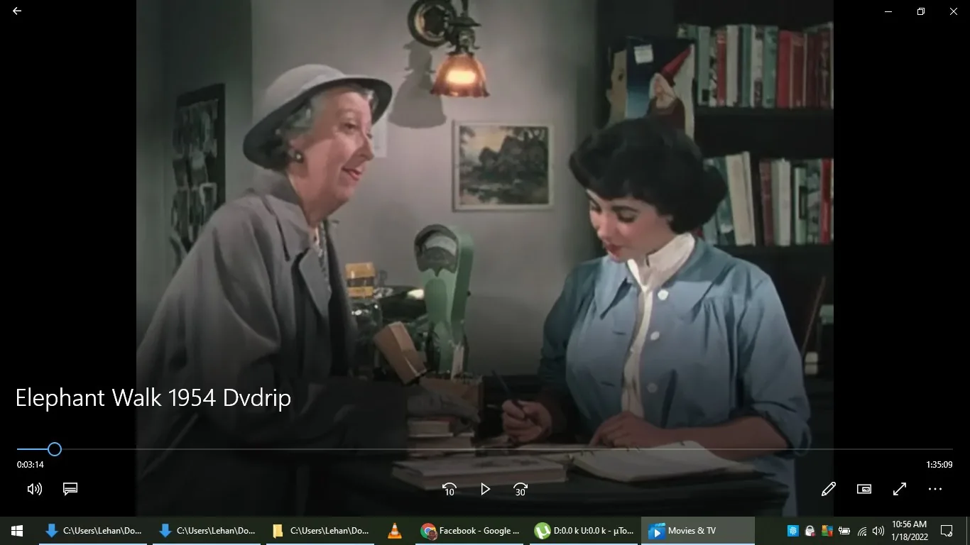 ```
The image is a video frame from a classic film titled "Elephant Walk" from 1954, as indicated by the on-screen text. The setting appears to be an indoor office or library, evidenced by the bookshelves filled with books in the background. Two women are engaged in a conversation. The woman on the left, wearing a gray coat and a hat, seems to be speaking, while the woman on the right, dressed in a blue button-up blouse, is writing something down in a notebook. The lighting is warm, coming from a lamp mounted on the wall in the background, which casts a soft glow over the scene. The decor includes a painting and a figurine, adding a touch of personality to the setting. The mood appears relaxed and professional, as they are focused on their tasks. The video appears to be playing in a media application, "Movies & TV," as suggested by the interface at the bottom of the frame. The scene is devoid of captions or subtitles, focusing attention on the interaction between the characters.