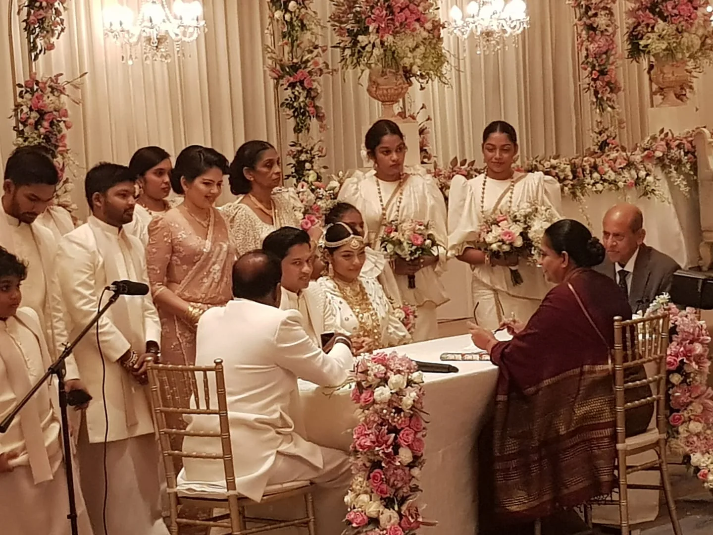 The image captures a formal wedding ceremony scene indoors, featuring a group of people gathered around a table adorned with floral arrangements. At the center of the scene, the bride and groom are seated, with the bride wearing an elegant white dress and ornate jewelry, indicative of a traditional wedding. The groom is dressed in a white suit. Surrounding the couple are bridesmaids in matching white attire, holding bouquets of flowers, and additional guests dressed in both traditional and formal wear.
The setting is elaborately decorated with flowers, and chandeliers hang from the ceiling, adding a luxurious touch to the ambiance. The people in the room, including Udhantha Sumathipala and Gamini Marapana, as identified by the hint, appear engaged in a moment of significance, likely involving the signing of wedding documents, as indicated by the presence of a table and a woman seated across with papers. The mood is formal and celebratory, with a harmonious blend of white and pastel colors complementing the overall decor.