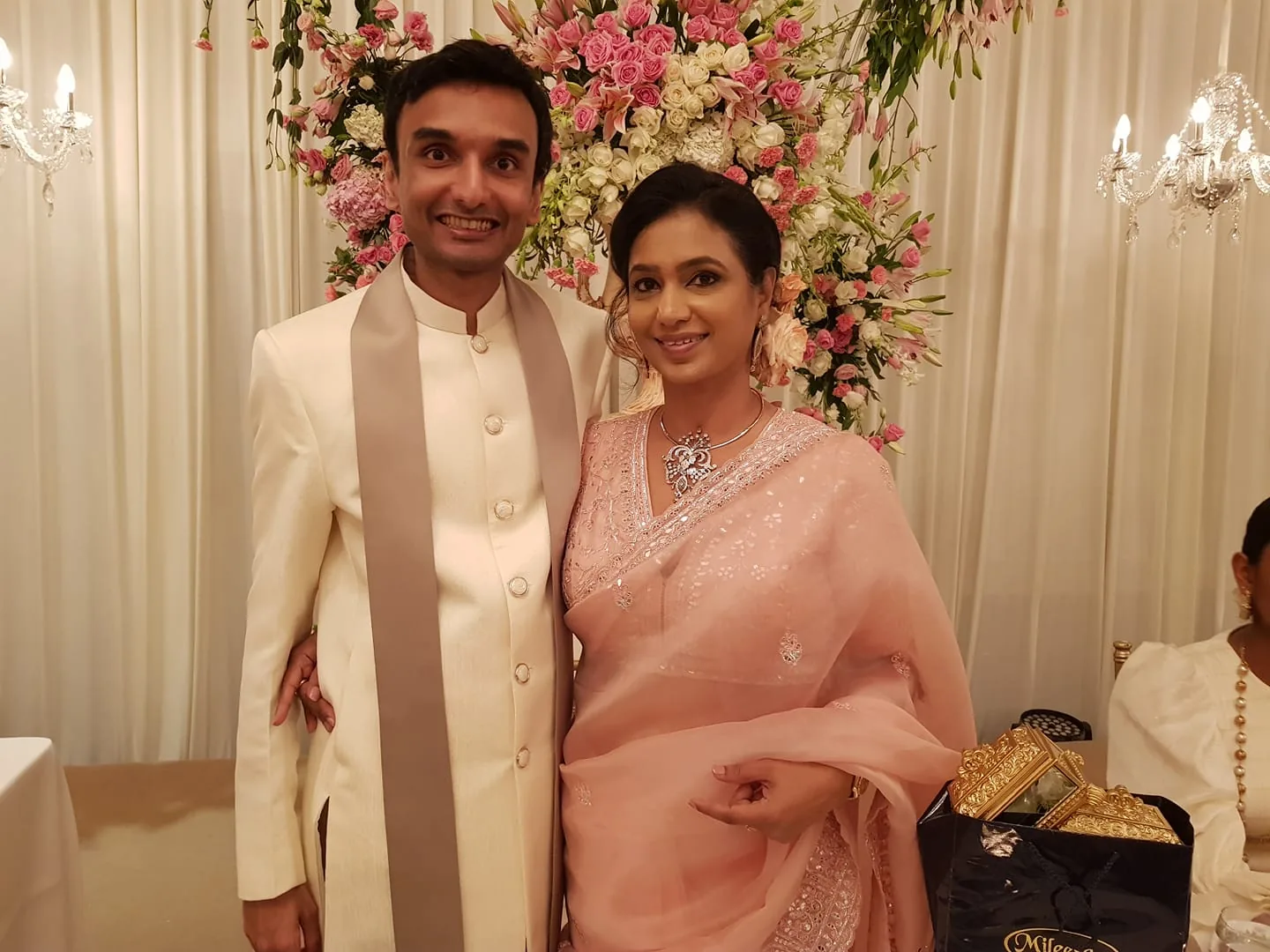 The image shows Lehan Edirisinghe and Shanika Edirisinghe in a formal setting, likely a wedding or similar celebration. They are standing closely together, smiling warmly at the camera. Lehan is wearing an elegant off-white sherwani with a light beige stole draped around his neck, exuding a sophisticated and traditional look. Shanika is adorned in a beautiful light pink saree with delicate embroidery and matching jewelry, including a necklace and earrings, complementing her attire.
Behind them is a lush floral arrangement with pink and white flowers, adding a vibrant and festive backdrop to the scene. The setting is indoors, with cream-colored drapes and elegant chandeliers enhancing the luxurious atmosphere. A bag labeled "Milestone" filled with ornate gold-colored objects is visible to the side. The mood is joyous and celebratory, capturing a moment of happiness and elegance.