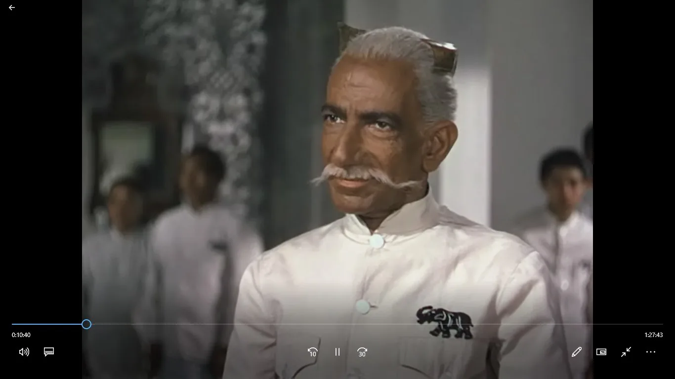 The image is a video frame from the 1954 film "Elephant Walk." It features an elderly man standing indoors, wearing a traditional white outfit with a high collar. The attire includes an embroidered elephant motif on his chest, adding a cultural touch to his formal appearance. His hair is white and neatly combed back, and he sports a prominent mustache, giving him a distinguished look. The background is slightly out of focus, with several men in similar white attire, suggesting a formal or official setting. The man’s expression is serious yet composed, indicating he might be engaged in a significant conversation or event. The lighting is soft and even, highlighting his facial features and the details of his outfit. The overall composition directs attention to the central figure, emphasizing his role in the scene.