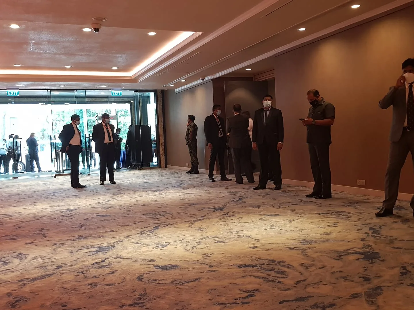 The image depicts an indoor scene in what appears to be a hotel lobby or conference area. The setting features a spacious carpeted floor with a patterned design in shades of blue and beige. A group of people, mostly men dressed in formal attire such as suits and ties, are scattered around the room. Some individuals are wearing masks, indicating a concern for health safety, possibly due to COVID-19 precautions.
The lighting is bright and comes from a combination of ceiling spotlights and natural light streaming through large glass doors in the background. The entrance is busy with people visible outside, suggesting an event or meeting is taking place. One person, possibly a security personnel, stands near the wall, distinctively dressed in a uniform. Another individual is using a phone, perhaps checking messages or making calls. The overall atmosphere is professional and somewhat subdued, with individuals appearing to be on standby or waiting.