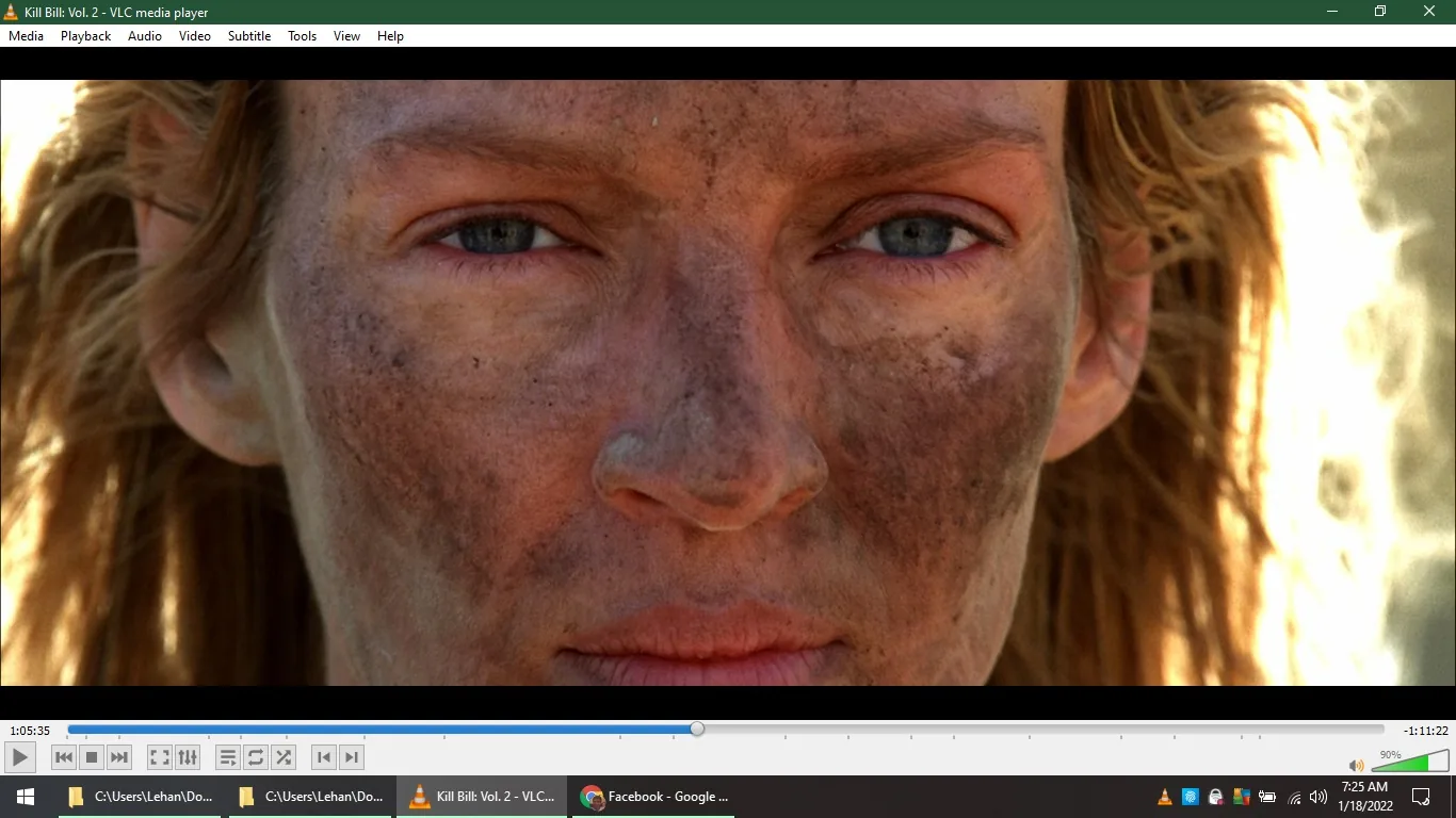 The image is a close-up shot from the film "Kill Bill: Vol. 2," viewed on VLC media player. It features Uma Thurman, who is the central focus. Her face is prominently displayed, covered in dirt and grime, suggesting a tense or action-packed sequence. The lighting is natural, highlighting her intense expression and the rugged texture of her skin. Her eyes are slightly narrowed, conveying determination and focus. The background is blurred, keeping the attention on her face. Her hair is loose and unkempt, further emphasizing the gritty and raw atmosphere of the scene. The VLC media player controls and time stamps are visible at the bottom, indicating the video's progress and playback settings. The timestamp shows the video is at 1 hour, 5 minutes, and 35 seconds with 1 hour and 11 minutes remaining, and the playback volume is at 90%. The computer's clock reads 7:25 AM on January 18, 2022.