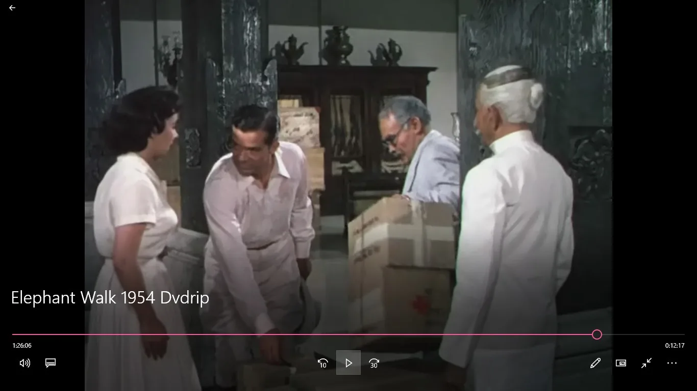The image is a frame from the 1954 film "Elephant Walk." It depicts a scene set indoors, possibly in a spacious room with ornate decor. In the foreground, a woman in a white dress is engaged in conversation with a man in a light-colored suit. He appears to be bending slightly, with his hand near a stack of boxes, suggesting they might be in the process of moving or organizing items. Two other men are present: one in a dark suit with glasses, and another in traditional white attire, likely indicating cultural or regional significance. The background shows a large wooden cabinet with decorative pieces on top, adding to the room's elegance. The lighting is bright, highlighting the characters and their actions. The attire and decor reflect the mid-20th century setting, contributing to the film's dramatic atmosphere. The title text on screen reads "Elephant Walk 1954 Dvdrip," indicating the film's name and format.