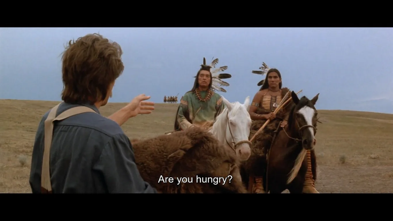 In this scene from the movie "Dances with Wolves," a man in the foreground, facing away from the camera, extends his hand towards two Native American men on horseback. The man's attire is rugged, typical of a frontier setting, with the landscape indicating a vast open prairie under a clear sky. The Native Americans wear traditional clothing adorned with feathers and beads, signifying their cultural attire. They sit on horses, one white and one brown, and the presence of a small group in the distance suggests a larger community. The lighting is natural, casting gentle shadows that enhance the earthy tones of the scene. The subtitle at the bottom reads, "Are you hungry?" indicating an offer or gesture of goodwill. The scene captures a moment of communication and potential understanding across cultural lines.