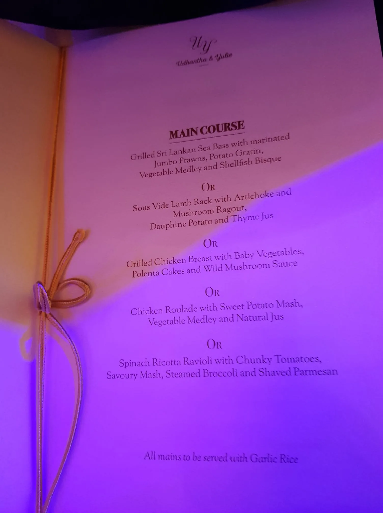 The image shows an elegant menu titled "Udhantha & Yukie," likely for a wedding or special event. The menu is presented on a page with a subtle, warm glow, possibly from ambient lighting or a nearby lamp. It's bound with a delicate golden string tied in a bow on the left side, adding a touch of sophistication. The main course options listed on the menu include a variety of gourmet dishes such as Grilled Sri Lankan Sea Bass, Sous Vide Lamb Rack, Grilled Chicken Breast, Chicken Roulade, and Spinach Ricotta Ravioli. Each dish is paired with an assortment of complementary sides, such as Potato Gratin, Mushroom Ragout, and Dauphine Potato. The menu notes that all main courses are accompanied by Garlic Rice. The text is elegantly printed, suggesting a formal and upscale dining experience. The use of purple lighting adds a modern and chic atmosphere to the setting.