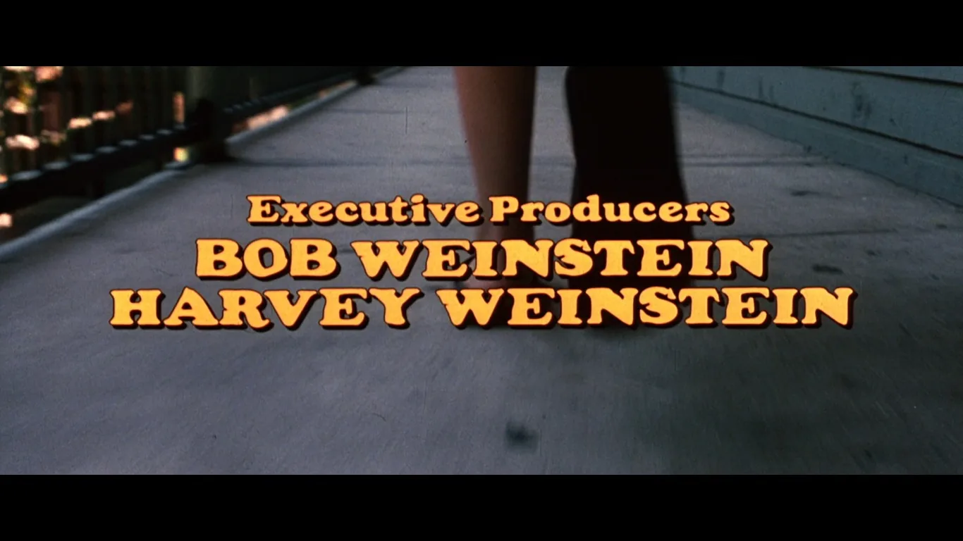 The image is a screenshot showing a credit sequence from a film or television production. The text overlay, centrally placed, reads "Executive Producers BOB WEINSTEIN HARVEY WEINSTEIN" in bold, yellow-orange lettering with a shadow effect, making it stand out against the background. The text is set on top of a blurred image of someone walking along a paved or wooden path with a railing on the left side, suggesting movement or transition. The lighting is natural, likely depicting an outdoor scene, with the background slightly out of focus to emphasize the text. The style of the text and the color palette give a retro or classic aesthetic, hinting at a film with that kind of tone. No specific site, app, or additional context is visible in the image.