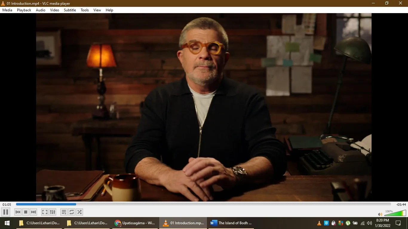 ```
1) Rich description: The image depicts a scene from a video titled "01 Introduction.mp4," played in VLC media player. The setting appears to be a cozy, wood-paneled room with a warm, ambient atmosphere. In the center, the person is presumably David Mamet, based on the provided hint. He is seated at a wooden desk, wearing a dark jacket over a white shirt. His facial expression is calm and composed, suggesting he is speaking or presenting. The desk is adorned with a few items, including a coffee mug and a stack of books, adding to the scholarly ambiance. Behind him, there is a softly lit table lamp contributing to the subdued lighting. On the wall, some notes or papers are pinned, enhancing the creative or intellectual vibe. The overall mood is intimate and focused, suitable for an introduction or discussion.
2) On-screen text block:
- Title: "01 Introduction.mp4"
- Site / app: "VLC media player"