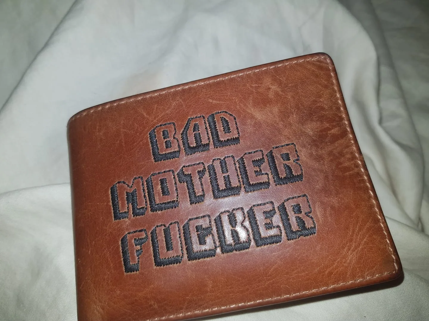 The image shows a close-up of a brown leather wallet lying on a white fabric surface, likely a bed or a couch. The wallet features prominently embroidered text in bold black letters that reads "BAD MOTHER FUCKER." The text is centered and stands out due to its contrasting color and the texture of the leather. The leather appears slightly worn, suggesting it might be well-used or intentionally distressed for aesthetic purposes. The stitching around the edges of the wallet is visible, and the overall lighting is even, providing a clear view of the texture and color of the materials. This style of wallet is reminiscent of a popular cultural reference, which adds a playful or bold statement to the item. The setting appears casual and relaxed, given the soft fabric background.