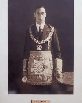 In this historical portrait, King George VI is depicted wearing formal Freemason regalia. The setting is a simple, indoor backdrop that highlights the solemnity and dignity of the occasion. King George VI stands upright, facing the camera, conveying a sense of authority and composure. He is dressed in a dark formal suit with a white dress shirt and a tie, over which he wears an elaborate Masonic apron featuring intricate embroidery and symbols, such as the square and compasses, and various other Masonic emblems. Around his neck, he wears a Masonic collar adorned with additional symbols and medals. The lighting is soft, casting subtle shadows that add depth to the image, while the sepia tone gives it a vintage, timeless quality. Below the portrait, there is a plaque with the text "KING GEORGE VI," identifying him explicitly. The overall ambiance of the photograph is formal and respectful, reflecting the historical significance of the subject.