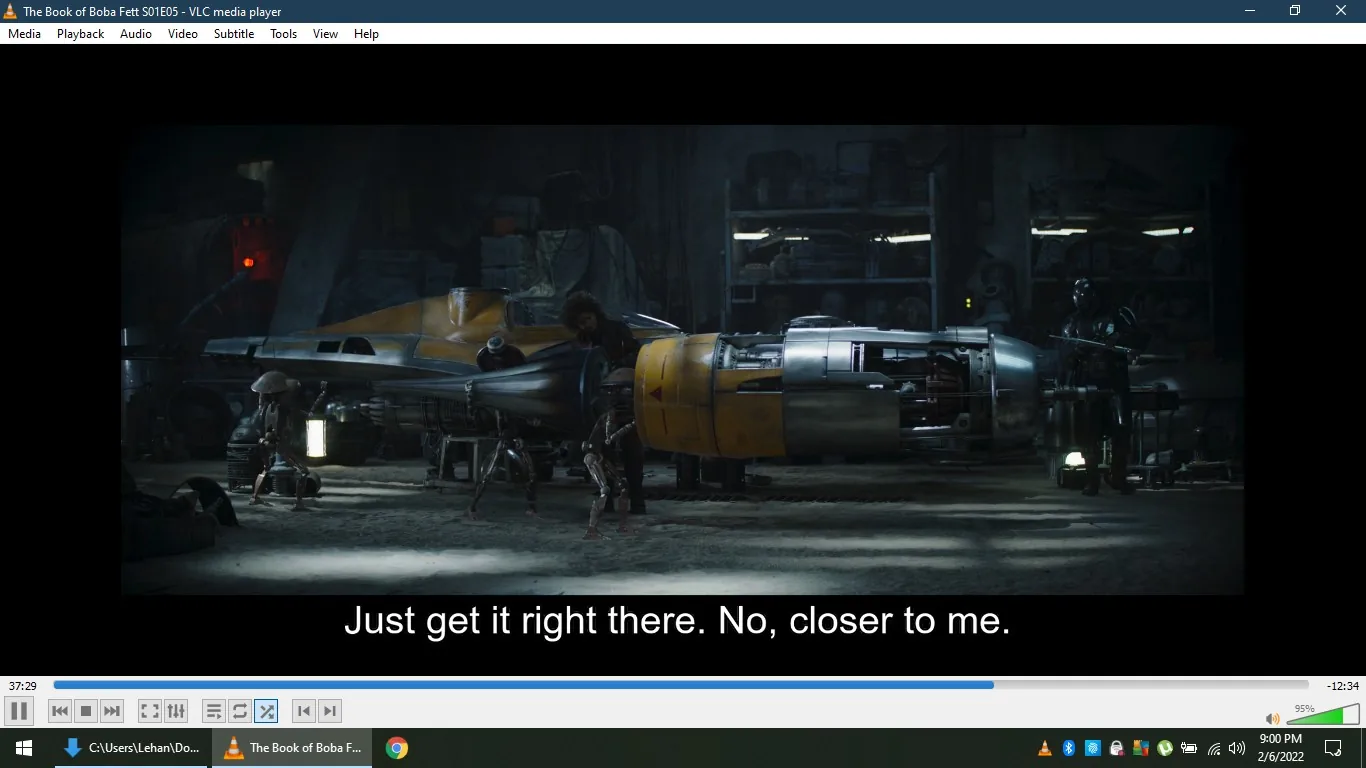 The image is a screenshot from "The Book of Boba Fett," specifically season 1, episode 5, as indicated by the title text in the VLC media player window. The scene takes place in a dimly lit workshop, featuring a partially assembled spacecraft that resembles a Naboo N-1 Starfighter. A character, possibly a mechanic, is working on the ship, assisted by a mechanical droid. Another individual, wearing dark armor typical of the Star Wars universe, stands nearby, observing the process. The lighting is moody, with shadows and focused light highlighting the spacecraft's metallic surfaces. The workshop is cluttered with various tools and equipment in the background. The subtitle reads, "Just get it right there. No, closer to me," indicating a precise task being carried out. The overall atmosphere suggests a scene focused on repair or modification, common in the Star Wars series.