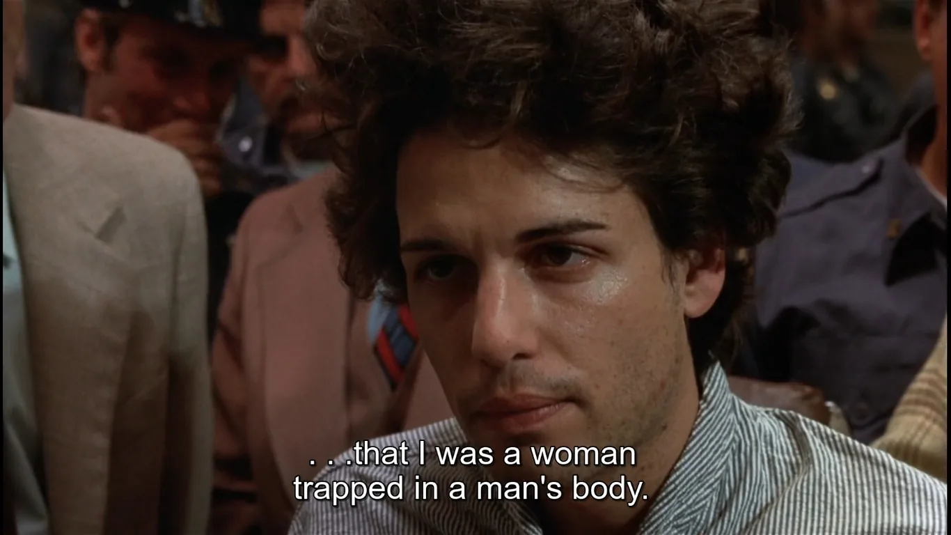 ```
The image is a frame from a movie scene set indoors, depicting a serious and tense atmosphere. The focal point is a person with curly hair, sitting among a group of people, possibly at a hearing or meeting. The person is wearing a striped shirt and has a contemplative expression, suggesting they are involved in a deep or revealing conversation. The background is filled with other people, some in uniforms, indicating a formal or official setting. The lighting is natural, highlighting the central figure's face, enhancing the emotional intensity of the moment. The subtitles at the bottom of the frame read, "...that I was a woman trapped in a man's body," indicating a discussion about identity and personal revelation. The scene captures an intimate and emotional moment, reflecting themes of gender identity and self-disclosure.