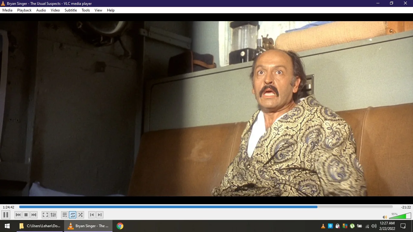 This image is a screenshot from a movie being played on VLC media player. The title at the top indicates it is from "The Usual Suspects," directed by Bryan Singer. The scene captures a man who appears to be sitting indoors, possibly in a train or a confined space due to the visible compartment and shelving. The man is wearing a patterned robe and has a surprised or startled expression on his face, with wide eyes and an open mouth. The lighting is somewhat dim, casting shadows on the wall behind him. The setting has a vintage feel, complemented by the muted colors and decor. The screenshot captures a moment of high tension or revelation, typical of the film's suspenseful style. The media player interface shows the timeline and controls at the bottom, indicating the video is near the end.
