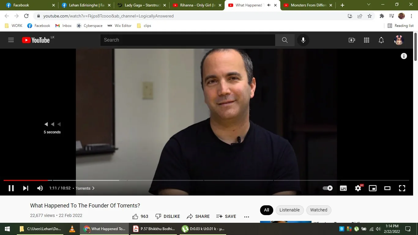 The image is a screenshot from a YouTube video titled "What Happened To The Founder Of Torrents?" from the channel LogicallyAnswered. The frame shows a man, referred to as Bram Cohen, seated indoors in what appears to be a casual interview setting. He is wearing a black t-shirt and has a microphone clipped to his shirt, suggesting he is speaking or being interviewed. The background is simple, with a plain wall and some dim lighting, focusing attention on the subject. Bram Cohen is looking at the camera with a slight smile, creating a relaxed and approachable atmosphere. The YouTube interface is visible, showing controls and video information, like the number of views and upload date. The scene is likely part of a discussion about torrents and their history, given the video's title and context.