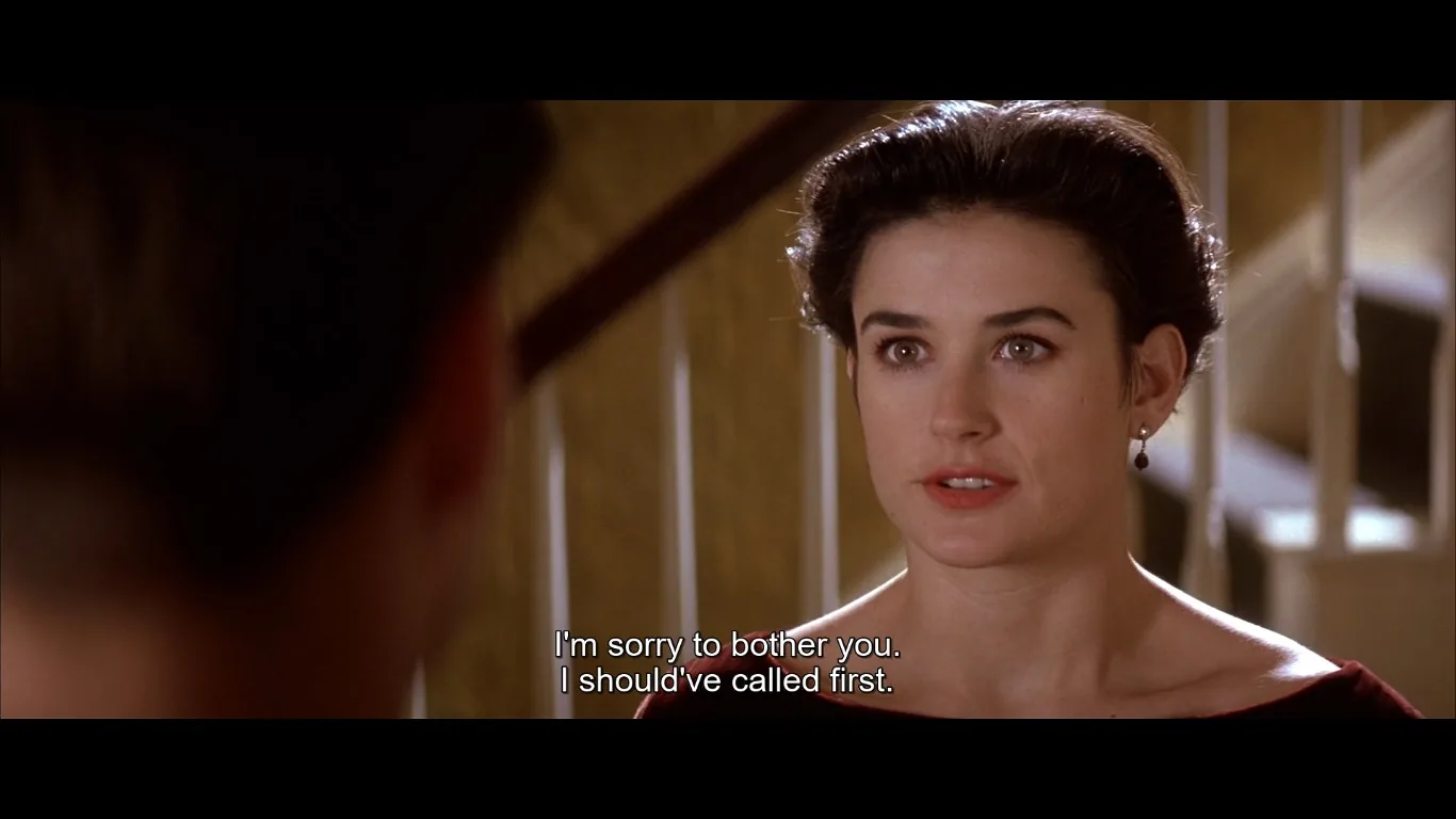The image shows a scene featuring Demi Moore, set indoors next to a staircase. The lighting is warm, suggesting a cozy or intimate environment. Demi Moore is positioned to the right of the frame, facing another person whose back is partially visible in the foreground, indicating a conversation. Her expression is earnest, and she is wearing simple earrings and a deep red top with an off-shoulder style. The composition centers on her facial expression, conveying sincerity. The background includes parts of a staircase, adding depth to the setting. The subtitles reveal her saying, "I'm sorry to bother you. I should've called first," which suggests a polite or apologetic tone in the interaction. The scene captures a moment of personal interaction, emphasizing dialogue and emotion.