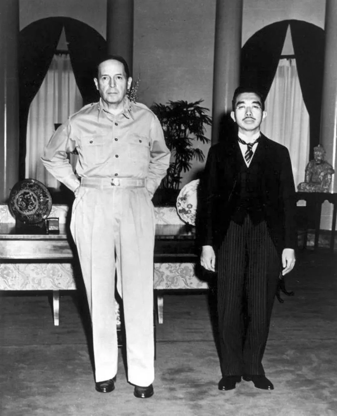 Hirohito and Douglas MacArthur are shown in a historical black-and-white photograph, symbolizing a significant post-World War II meeting. Both figures are standing side by side in a formal setting. Hirohito, wearing a dark formal suit with a tie, appears solemn and composed. His attire is traditional, with a striped pattern visible on his trousers, and he stands with his hands at his sides. Douglas MacArthur, dressed in a light-colored military uniform, exudes a commanding presence. His posture is confident, with hands behind his back. The background features elegant decor, including ornate plates on a table, lush plants, and statues, suggesting a grand and official venue. The lighting is soft, emphasizing the serious and formal nature of the meeting, which historically marked a turning point in the relationship between Japan and the United States during the occupation period.