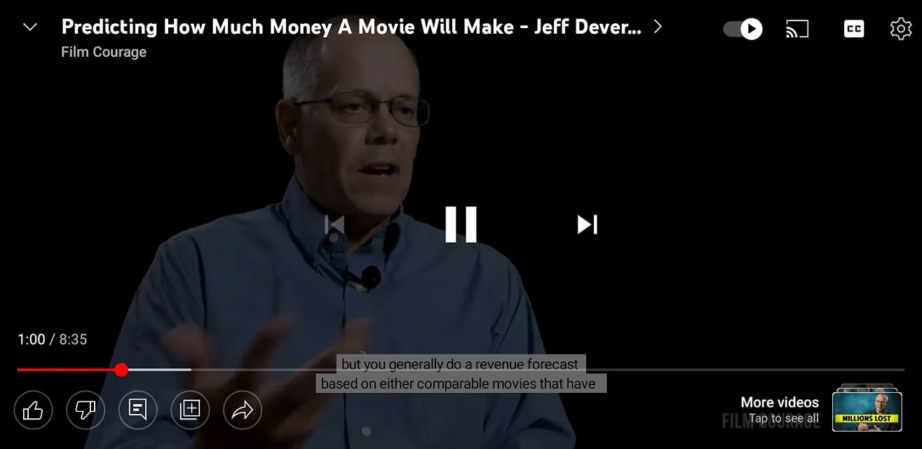 The image is a video frame from a YouTube interview titled "Predicting How Much Money A Movie Will Make - Jeff Deverett" on the Film Courage channel. The scene features Jeff Deverett in an interview setting, likely discussing aspects of the film industry's financial forecasting. Jeff is wearing glasses and a blue button-up shirt, with a small microphone clipped to his collar, suggesting he is speaking or being interviewed. The background is dark, focusing attention on Jeff, who is the central figure. The video is paused at the 1:00 mark out of a total duration of 8:35, as indicated by the progress bar at the bottom. Captions are visible, with Jeff speaking about revenue forecasting based on comparable movies. The lighting highlights Jeff against the dark backdrop, emphasizing the serious, informative mood of the interview.