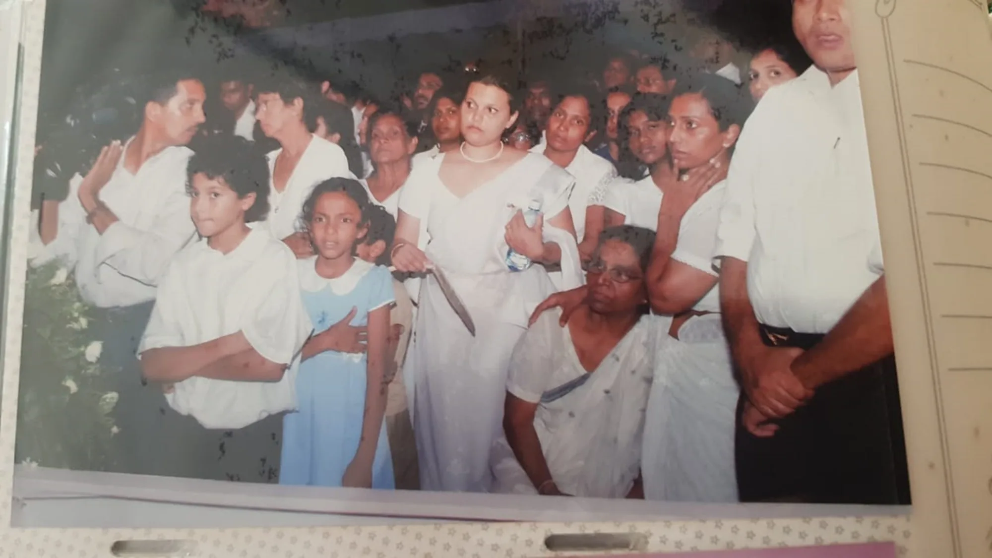 The image captures a group of people gathered closely together, suggesting a formal or solemn event. The setting appears to be indoors, possibly at a ceremony or memorial, as evidenced by the expressions and attire of the attendees. Among the crowd, a woman dressed in a white sari stands out, holding a bottle of water and a fan, indicative of a warm environment. The people around her, including children and adults, are dressed in formal attire, with the children wearing a white shirt and a blue dress, respectively.
The individuals in the image convey a range of emotions from solemnity to contemplation. The woman in the sari, likely Manjaree Gamage, has a pensive expression, while the older woman seated in front, possibly Milina Sumathipala, appears reflective. A man in the crowd, potentially Mikey de Alwis, is seen holding a camera to his shoulder, suggesting documentation of the event. The lighting is subdued, emphasizing the serious or respectful nature of the gathering. Overall, the image suggests a community coming together, possibly in remembrance or support.