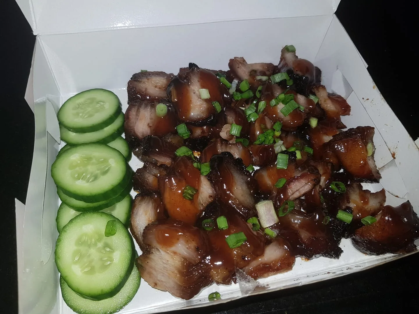 The image displays a takeout meal consisting of sliced roasted pork topped with a glossy, dark sauce and garnished with chopped green onions. The pork appears caramelized and juicy, indicating it might be char siu, a popular Chinese-style barbecue dish. Next to the meat, there are several slices of fresh cucumber, providing a contrasting green color and crunchy texture to the dish. The food is placed in a white takeout box, suggesting it might be from a restaurant or delivery service, designed for easy transport and consumption. The lighting is bright, highlighting the sheen of the sauce and the texture of the pork. The overall presentation is neat, with the cucumbers arranged in a tidy stack, enhancing the visual appeal of the meal. There are no visible logos or text in the image to indicate a specific restaurant or brand.