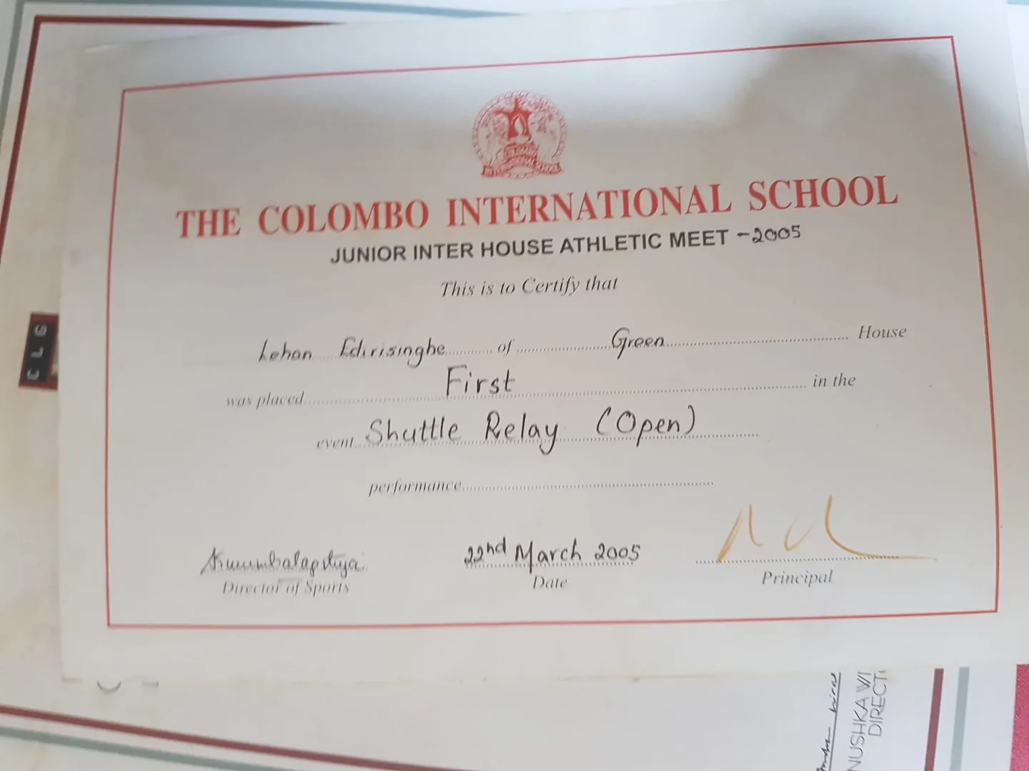 This image shows a certificate awarded by The Colombo International School for a Junior Inter House Athletic Meet in 2005. The certificate is primarily in white with a red border and text. At the top, the institution's name is prominently displayed in bold red letters: "THE COLOMBO INTERNATIONAL SCHOOL." Below, the certificate details indicate it is for a first-place finish in the Shuttle Relay (Open) event. Specific fields include placeholders for the recipient's name and house, which appear to be filled in with handwritten text. The document is signed by the Director of Sports and the Principal, with the date noted as 22nd March 2005. The certificate is likely printed on a standard sheet of paper, possibly framed or displayed against another document in the background. The overall composition suggests an official and formal recognition of athletic achievement within the school setting.