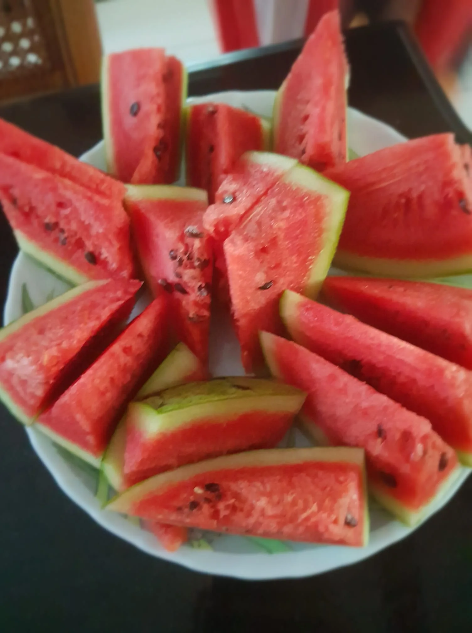 The image features a close-up view of a plate filled with neatly arranged slices of watermelon. The slices are triangular, showcasing the vibrant red flesh and contrasting light green rind. Each piece is positioned upright, creating a visually appealing pattern in the dish. The watermelon seeds are visible, adding texture to the fruit's surface. The setting appears to be indoors, possibly on a dining table, as indicated by the dark surface underneath the plate. The lighting is soft, highlighting the juicy and fresh appearance of the watermelon. The overall mood of the image is refreshing and summery, evoking a sense of freshness and thirst-quenching appeal. No visible text or branding is present. The composition emphasizes the natural beauty and color of the fruit, making it an inviting treat.