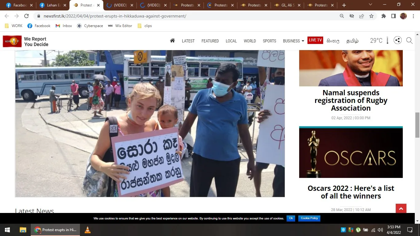 The image is a screenshot from the website "newsfirst.lk," showing a scene from a protest in Hikkaduwa, Sri Lanka, against the government. In the foreground, a woman holding a child is prominently displaying a sign written in Sinhala. She appears to be participating in the protest, suggesting determination and resolve. Next to her, a man wearing a blue shirt and mask also holds a protest sign, indicating active involvement in the demonstration. The setting is outdoors, with buses and people visible in the background, reinforcing the public nature of the event. The lighting is bright, indicating it is daytime. On the right side of the screen, there are headlines about Namal suspending the registration of the Rugby Association and a list of Oscar winners for 2022, reflecting other news topics covered by the website. The overall mood of the image conveys activism and civic engagement.
- Title: "Protest erupts in Hikkaduwa against government"
- Site / app: "newsfirst.lk"