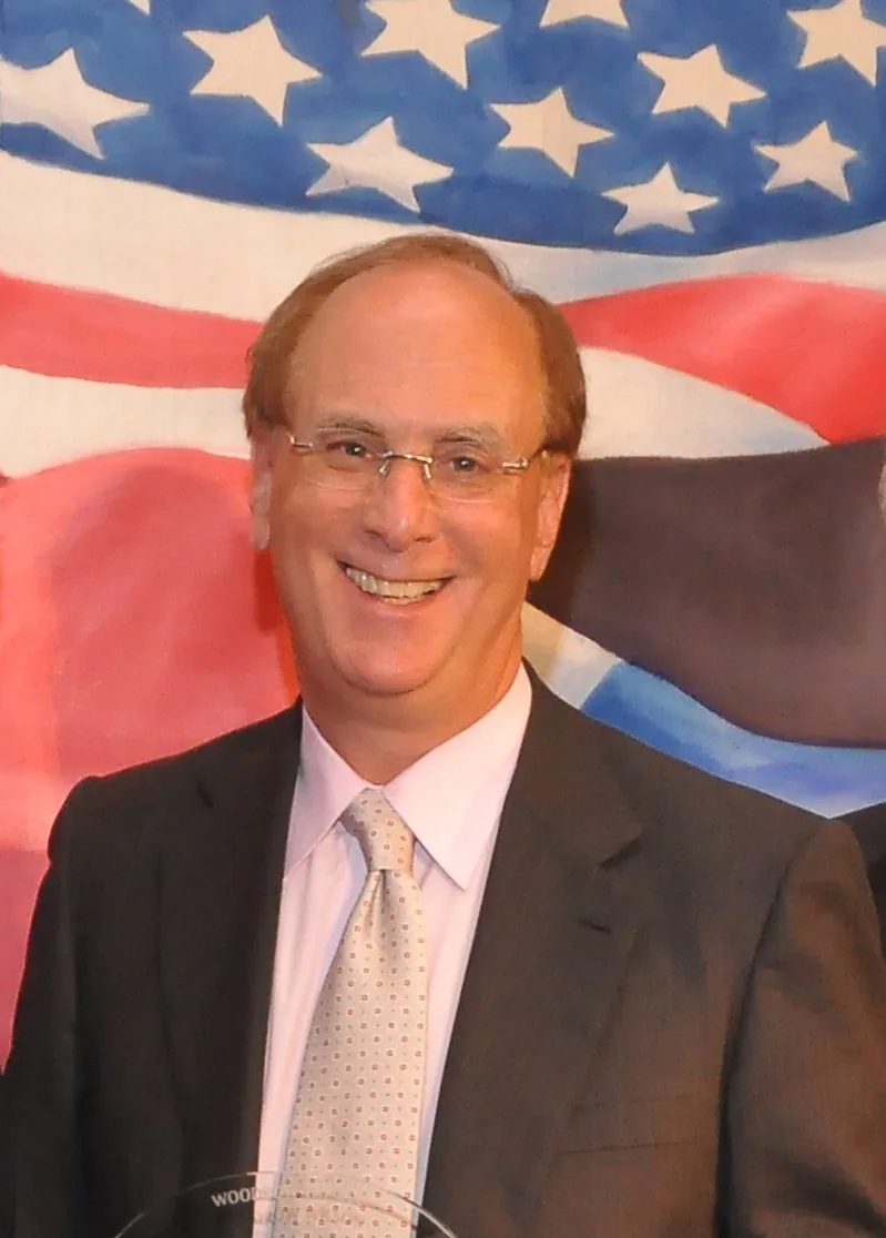 The image features Lawrence Fink, associated with BlackRock, in a formal setting. He is wearing a dark suit paired with a light pink shirt and a polka-dotted tie, suggesting a professional event or gathering. His expression is cheerful, and he seems to be in a positive mood, possibly celebrating an achievement or attending a significant event. The background prominently displays an American flag with stars and stripes, which adds to the formal and patriotic atmosphere of the scene. The lighting is bright and even, emphasizing a clear and crisp image, typical for event photography. The composition focuses on Fink, highlighting him as the central figure against the flag backdrop, which further underscores the importance of the occasion.