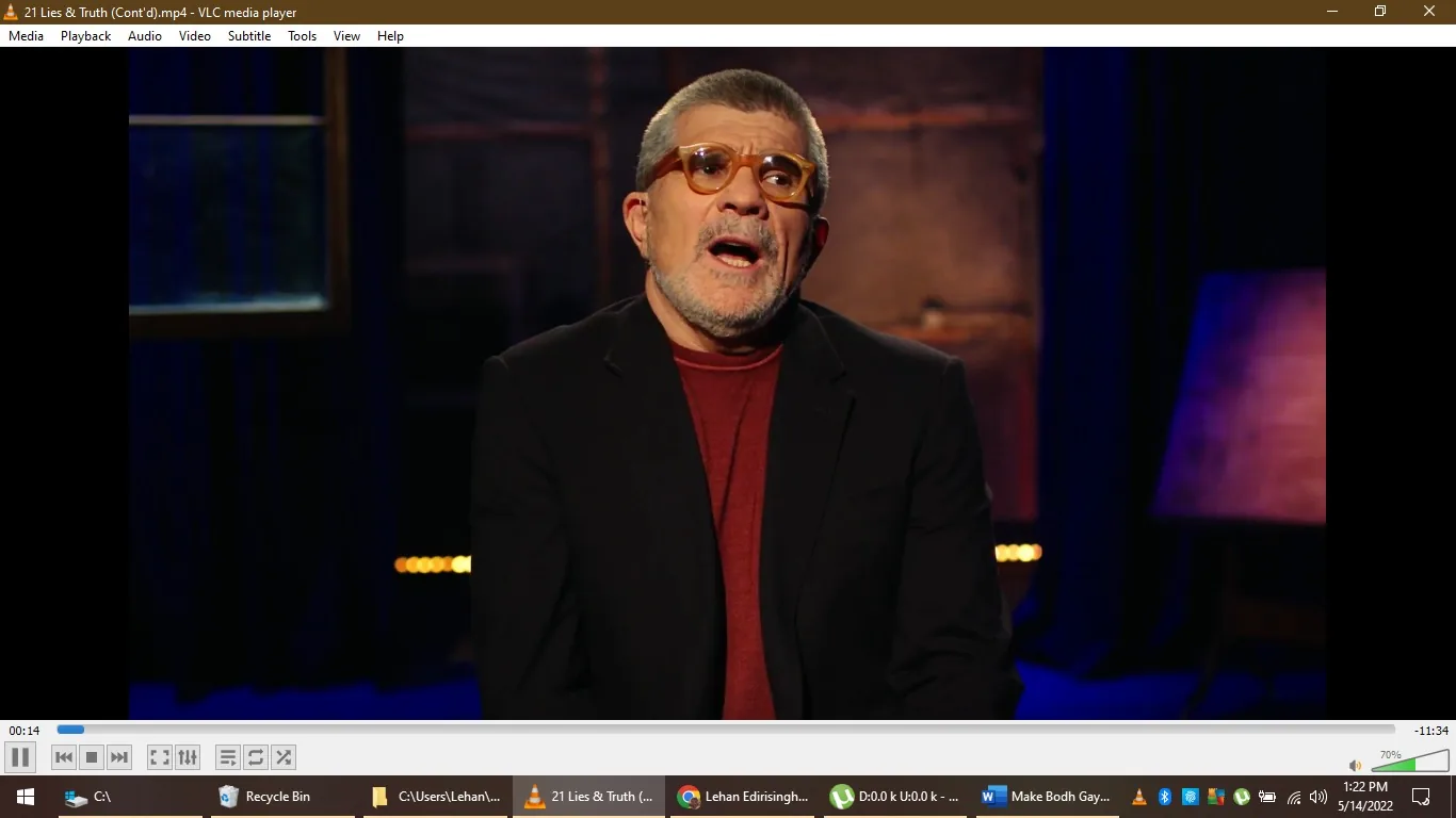 David Mamet is seen in this video frame from a file titled "21 Lies & Truth (Cont'd).mp4," played on VLC media player. He appears to be in an indoor studio setting, as indicated by the dark background with subtle lighting. Mamet is seated, wearing a black blazer over a red shirt, and he has short gray hair and a beard. The lighting is warm, highlighting his facial features and creating a focused ambiance typical of interview or discussion formats. His expression seems engaged, possibly as he speaks on a particular topic. The background is out of focus, with hints of blue and orange tones, adding to the studio atmosphere. The interface of VLC media player is visible, showing standard playback controls and a timeline indicating 14 seconds into an 11-minute and 34-second video. No captions or subtitles are present on the screen.