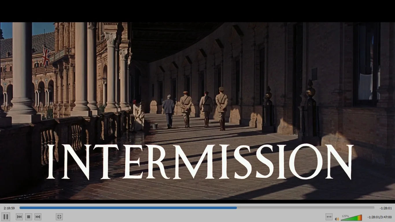 In this video frame from the film "Lawrence of Arabia," the scene is set in an expansive outdoor corridor of a historic building, likely representing a location in the film's Middle Eastern setting. The architecture features grand columns and arches, exhibiting a classical design with intricate details. Several individuals, dressed in period military uniforms and civilian attire, are walking through the corridor, casting long shadows on the tiled floor due to the bright sunlight. The atmosphere conveys a sense of historical grandeur and military formality. A British flag can be faintly seen in the background, hinting at the colonial context of the setting. The word "INTERMISSION" is prominently displayed across the bottom of the image, indicating a break in the film's narrative. The lighting is warm, contributing to the majestic and timeless feel of the scene, fitting the epic nature of the film.