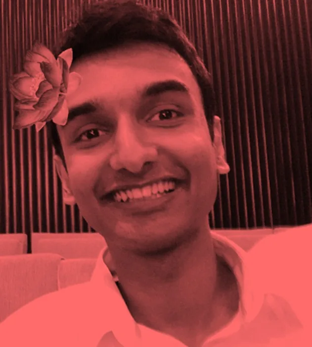 The image features a portrait of a person identified as Lehan Edirisinghe. The photo is stylized with a red monochrome filter, giving it a distinct and artistic look. Lehan is smiling, creating a warm and friendly atmosphere. A digital flower accessory is placed on the left side of his head, adding a playful and whimsical element to the composition. He is wearing a light-colored shirt, and the background consists of vertical lines, possibly wood paneling, suggesting an indoor setting like a theater or auditorium. The overall mood is casual and cheerful, aligning with the lighthearted nature of the flower accessory. This digitally enhanced portrait highlights a combination of natural expression and creative editing.