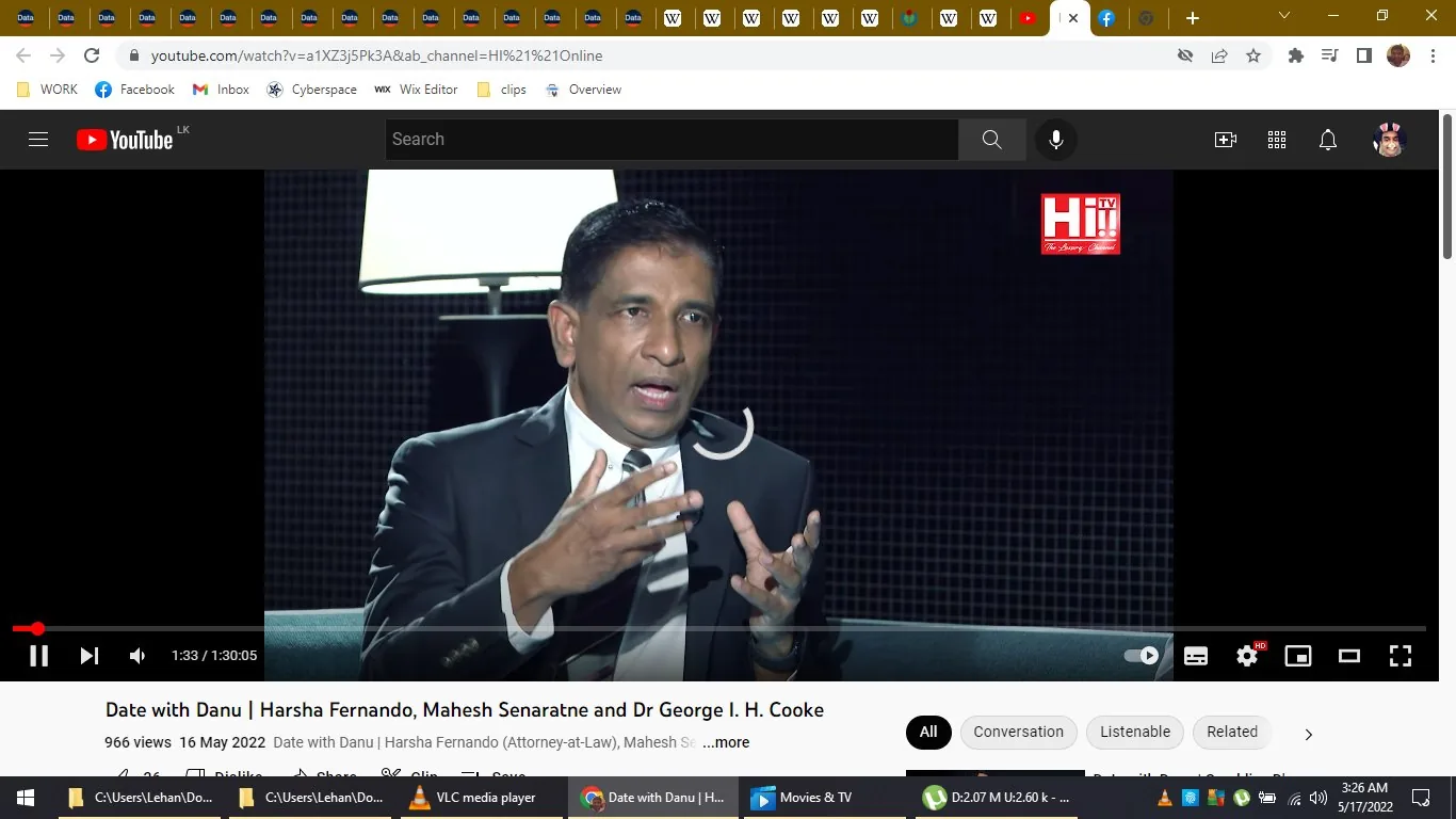 The image is a video frame from a YouTube video titled "Date with Danu | Harsha Fernando, Mahesh Senaratne and Dr George I. H. Cooke" on the channel HI!! Online. The setting appears to be an indoor interview or talk show, as indicated by the presence of a person speaking while seated, dressed in formal attire—a dark suit and tie. The lighting is professional and focuses on the individual, suggesting a studio environment. The person is speaking animatedly, using hand gestures, indicating they are engaged in a discussion or explanation. In the background, a lamp and a textured backdrop create a formal, studio-like atmosphere. The branding for "HI!! The Luxury Channel" is visible on the upper right corner of the screen. The time stamp shows 1:33 out of a 1:30:05 long video, suggesting an early segment. The video appears to be a talk show or interview format, focusing on conversations with Harsha Fernando among other guests.