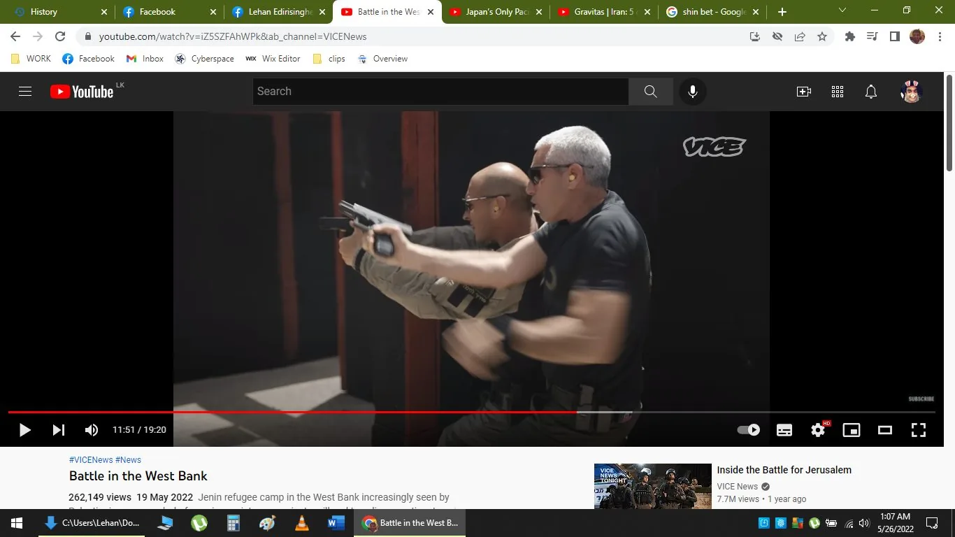 The image is a YouTube screenshot showing a scene from a video titled "Battle in the West Bank," published by VICE News. The video is paused at 11:51 out of a total 19:20 duration. Two individuals are in an indoor setting, seemingly involved in firearms training. Both are wearing sunglasses and black clothing, indicating a tactical or security context. The man in front is holding a gun, pointing forward, with a serious and focused demeanor. The lighting is bright, emphasizing the serious mood of the scene. The video, published on May 19, 2022, discusses the Jenin refugee camp in the West Bank. The YouTube interface shows tabs and bookmarks at the top, including Facebook and other YouTube videos, highlighting a typical browsing environment.
- Title: "Battle in the West Bank"
- Channel / profile: "VICE News"
- Site / app: "YouTube"