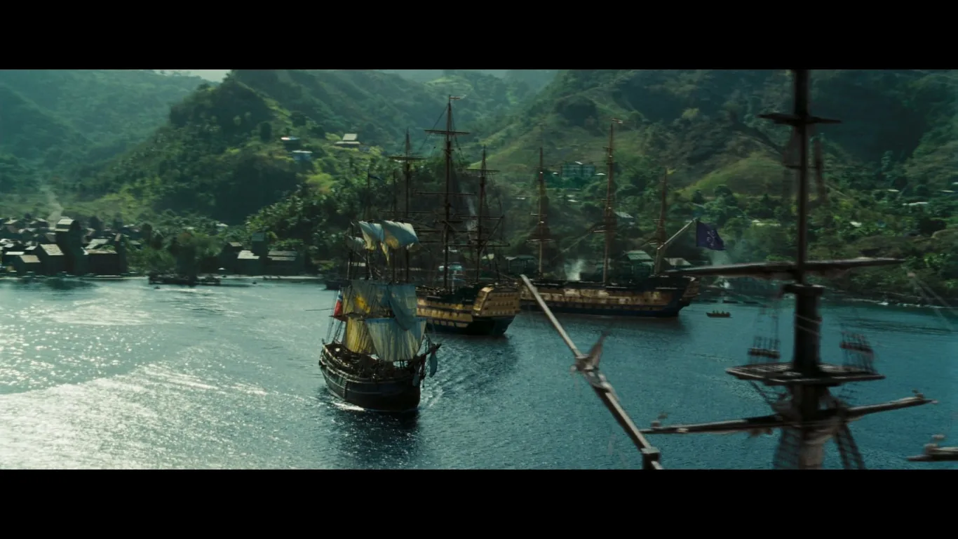 The image captures a picturesque maritime scene featuring several grand sailing ships anchored in a scenic harbor, likely from the "Pirates of the Caribbean" series. The foremost ship is adorned with worn sails, indicating wear and tear from long voyages. Behind it, other ships are visible, including one flying a pirate flag, suggesting a setting related to piracy. The water reflects the bright sunlight, creating a shimmering effect. In the background, a lush, mountainous landscape stretches across the horizon, dotted with what appear to be small buildings or a village nestled along the coastline. The atmosphere conveys a sense of adventure and exploration typical of the "Pirates of the Caribbean" films, with the presence of ships and natural beauty. The overall lighting is bright, enhancing the vivid colors of the scenery.