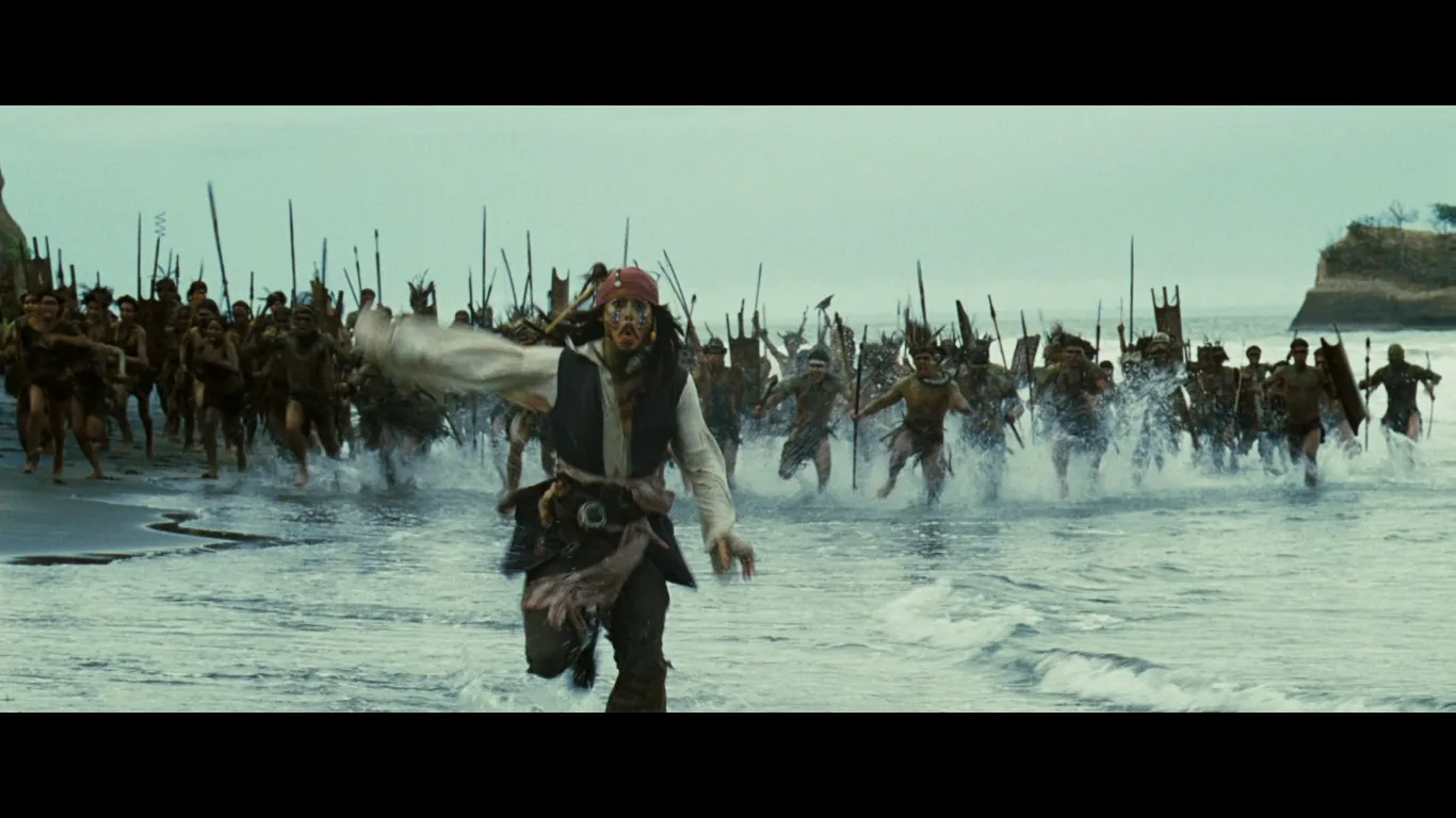 ```
In this dynamic scene from "Pirates of the Caribbean," Jack Sparrow is shown frantically running along a beach. He is pursued by a large group of islanders wielding spears and makeshift weapons, charging through shallow water. Jack Sparrow, characterized by his iconic pirate attire, including a red bandana, a vest, and various accessories hanging from his belt, appears in the foreground. His expression conveys urgency and panic as he glances backward at his pursuers. The background is a vast expanse of ocean and sky, with the islanders forming a cohesive line of pursuit. The lighting is natural, suggesting daytime, and the overall mood is one of intense, chaotic action. The scene's composition emphasizes the sense of speed and urgency as Jack desperately tries to escape.