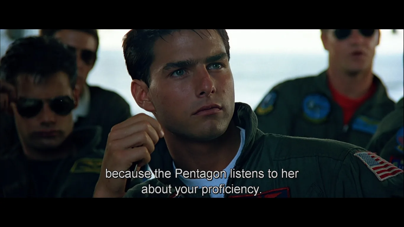 ```
In this scene from the film "Top Gun," the focus is on Tom Cruise's character, who appears engaged in a serious military briefing. He is dressed in a green flight suit adorned with patches, including an American flag on his shoulder, indicating his role as a pilot. The setting is a classroom or briefing room, with other pilots visible in the background, some wearing sunglasses, which adds to the intense and focused atmosphere.
The lighting highlights the expressions of the characters, particularly emphasizing the determined and attentive look on Tom Cruise's face. The mood is serious, reflecting the high-stakes environment of military aviation training. The subtitle reads, "because the Pentagon listens to her about your proficiency," suggesting a discussion of performance evaluation or a briefing on strategic assessments. The composition places Cruise in the foreground, making him the focal point of the scene, while other pilots are slightly out of focus, underscoring his central role in the narrative at this moment.