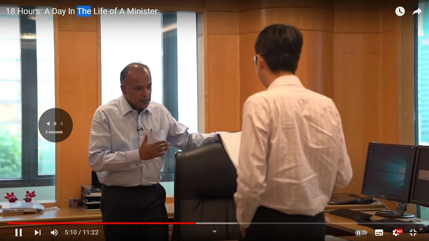 1) Rich description: The image is a video frame from a documentary titled "18 Hours: A Day In The Life of A Minister." It features K Shanmugam, who is likely engaged in a discussion within an office setting. He is wearing a light blue dress shirt, which is slightly open at the collar, suggesting a casual yet professional environment. Opposite him stands another individual in a white striped shirt, holding a notebook or document. The interaction appears serious and focused, with K Shanmugam gesturing with one hand, possibly emphasizing a point. The office is well-lit with ample natural light coming through large windows in the background, and the walls are paneled with wood, creating a warm and formal ambiance. There are computer monitors on a desk to the side, indicating a modern workspace. The overall atmosphere is one of a busy, professional setting typical of a high-level government or administrative office.
2) On-screen text block:
- Title: "18 Hours: A Day In The Life of A Minister"