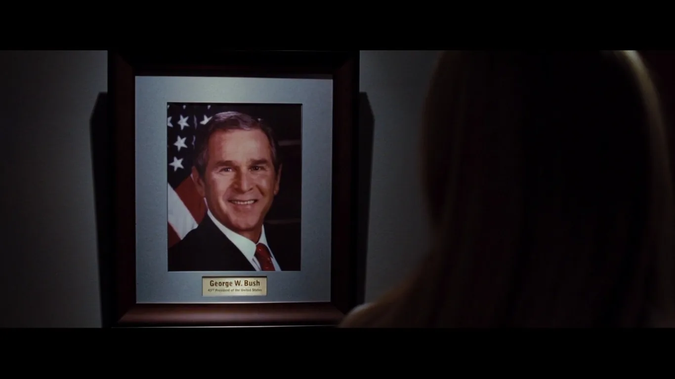 The image features a framed photograph of George W. Bush, identified as the 43rd President of the United States. The photograph depicts him smiling, and it is mounted against a backdrop that includes elements of the American flag, suggesting a presidential setting. The frame is simple with a dark border, and a plaque beneath the photograph clearly states his name and title. The lighting in the scene is subdued, highlighting the photograph while casting shadows on the wall. In the foreground, the back of a person's head is visible, suggesting they are observing the photograph. The overall mood is formal and respectful, typical of official portraits in governmental or historical contexts. The setting appears to be indoors, likely in a corridor or gallery where such portraits are displayed. The focus is entirely on the photograph of George W. Bush, indicating its significance in the scene.