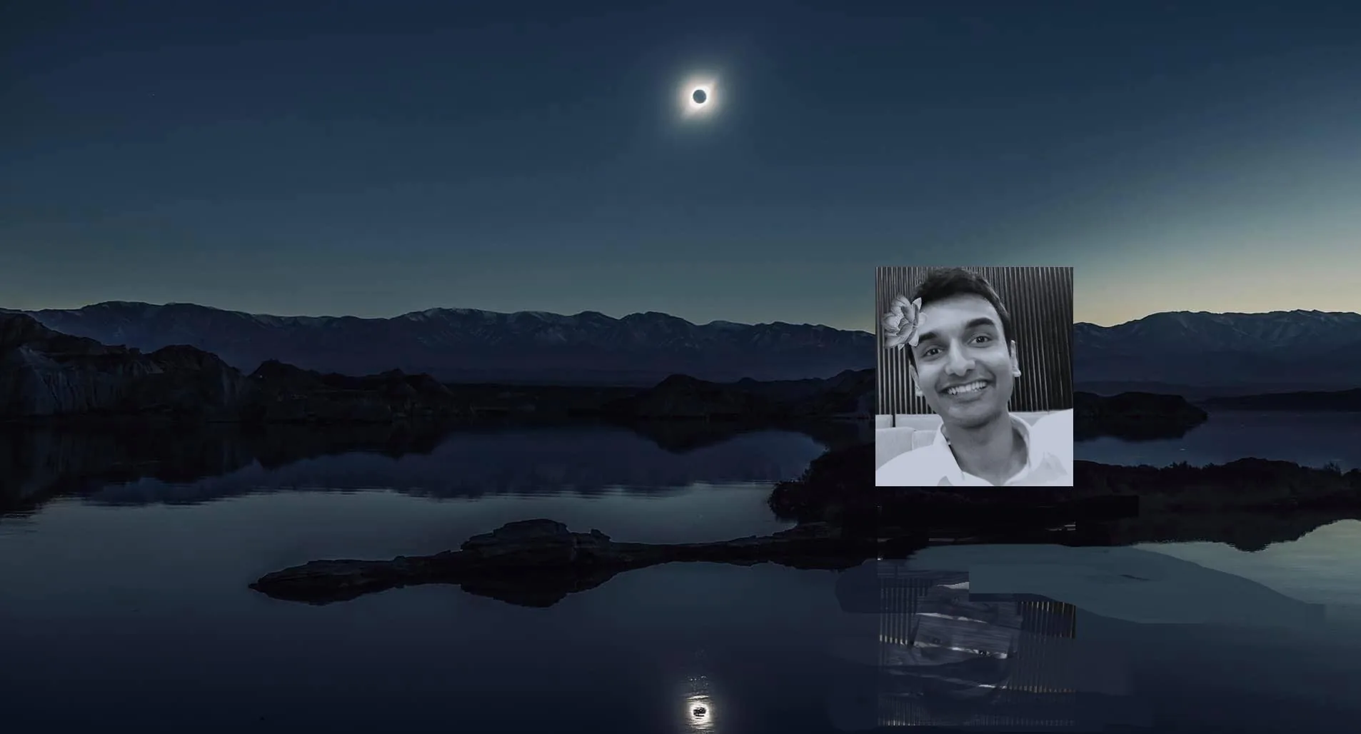 This image features a striking juxtaposition of a landscape and a portrait. The background shows a serene nighttime scene with a total solar eclipse occurring over a calm body of water. The moon is centered in the sky, casting a glow around its edges, creating an ethereal halo effect. The surrounding landscape consists of silhouetted mountains and their reflection in the water, contributing to the tranquil atmosphere.
In the foreground, there is a black-and-white portrait of a person, identified from the hint as Lehan Edirisinghe. The individual is smiling warmly and has a flower tucked behind one ear, adding a playful touch to the image. The background of the portrait appears to be indoors, with a patterned wall visible. The lighting in the portrait is soft, highlighting the person's facial features.
The composition contrasts the natural eclipse scene with the intimate, personal portrait, blending celestial and human elements in an intriguing manner. The overall tone of the image is serene and contemplative, with a mix of natural wonder and personal expression.